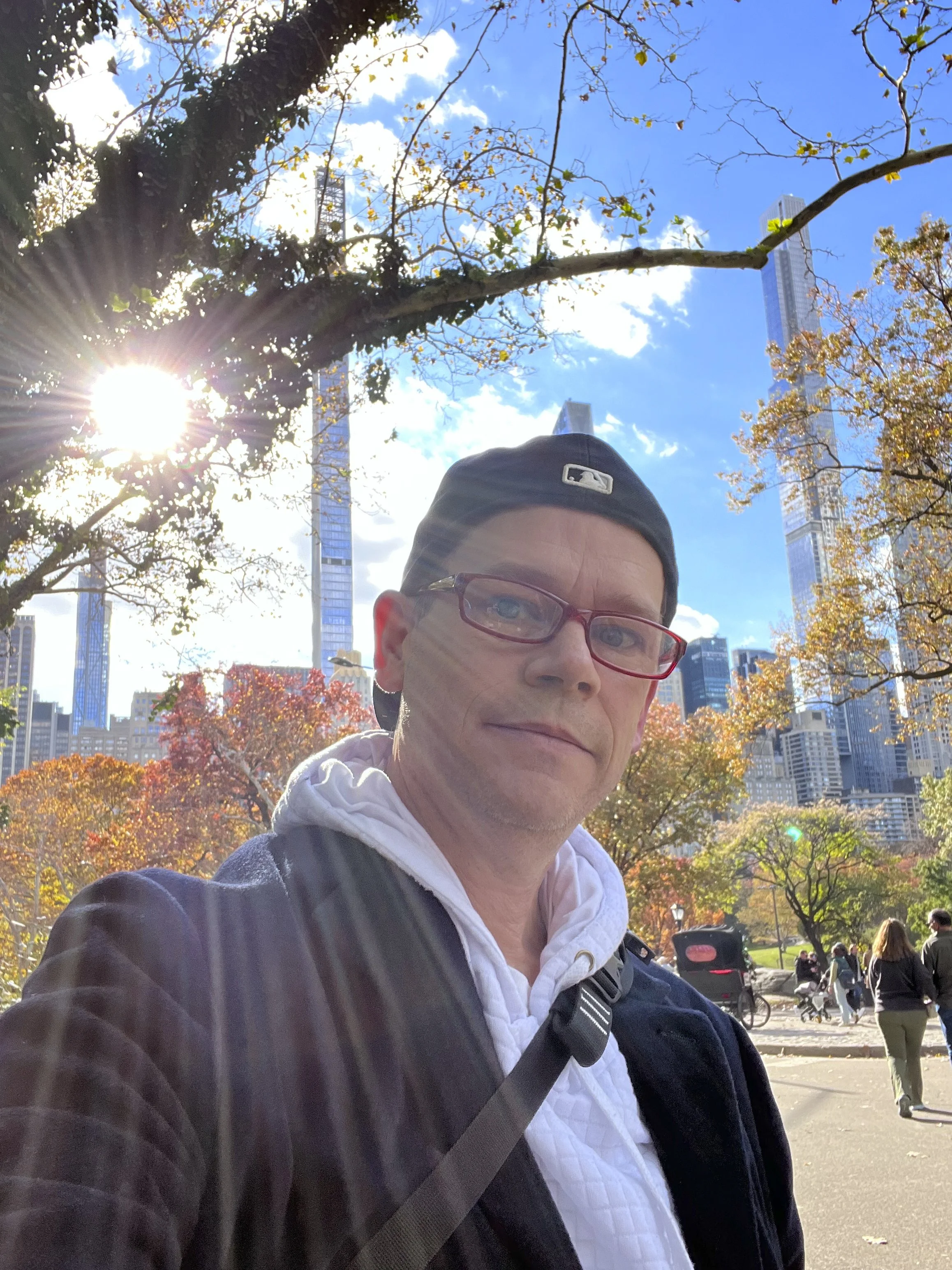 Ken taking a selfie in NYC's Central Park with fall foliage and tall buildings of the New York City skyline in the background, with the sun shining through the trees.