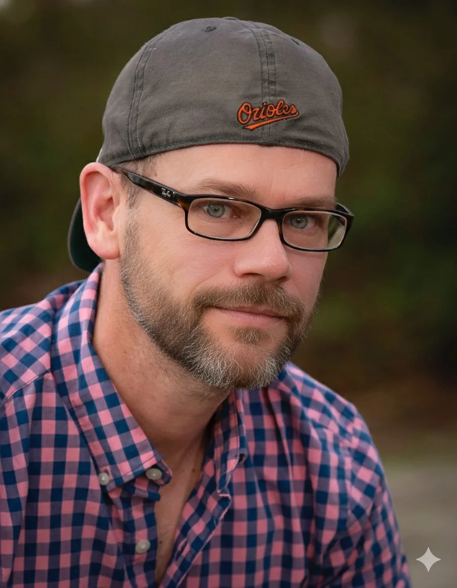 A man wearing glasses, a gray baseball cap with orange embroidery, and a pink and blue checkered button-up shirt.