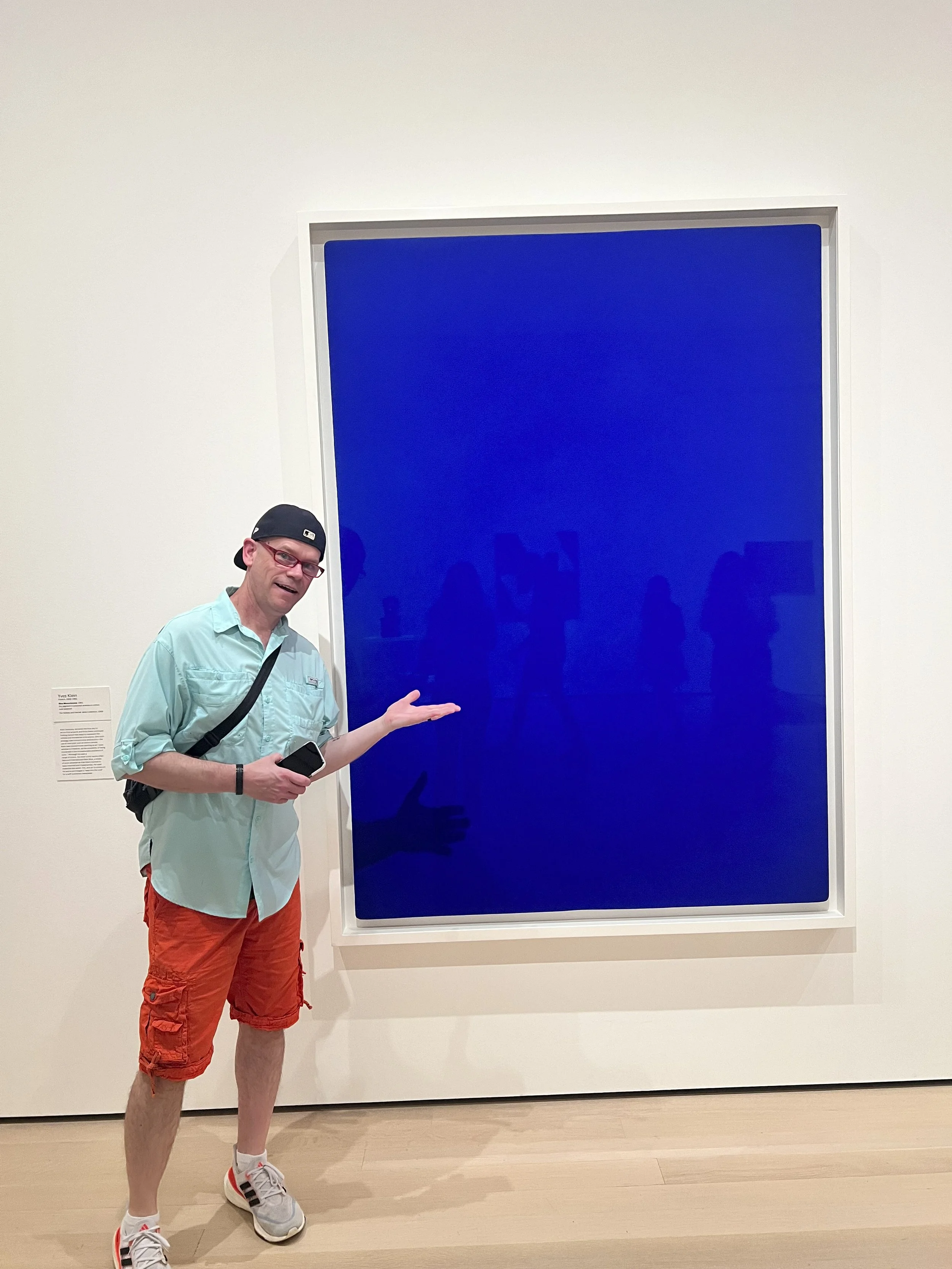 A man, Ken,  in a turquoise shirt, orange shorts, gray sneakers, and a black cap wearing glasses stands in front of a large, deep blue monochromatic painting in a gallery at the MET. He is gesturing towards the artwork while holding his phone.