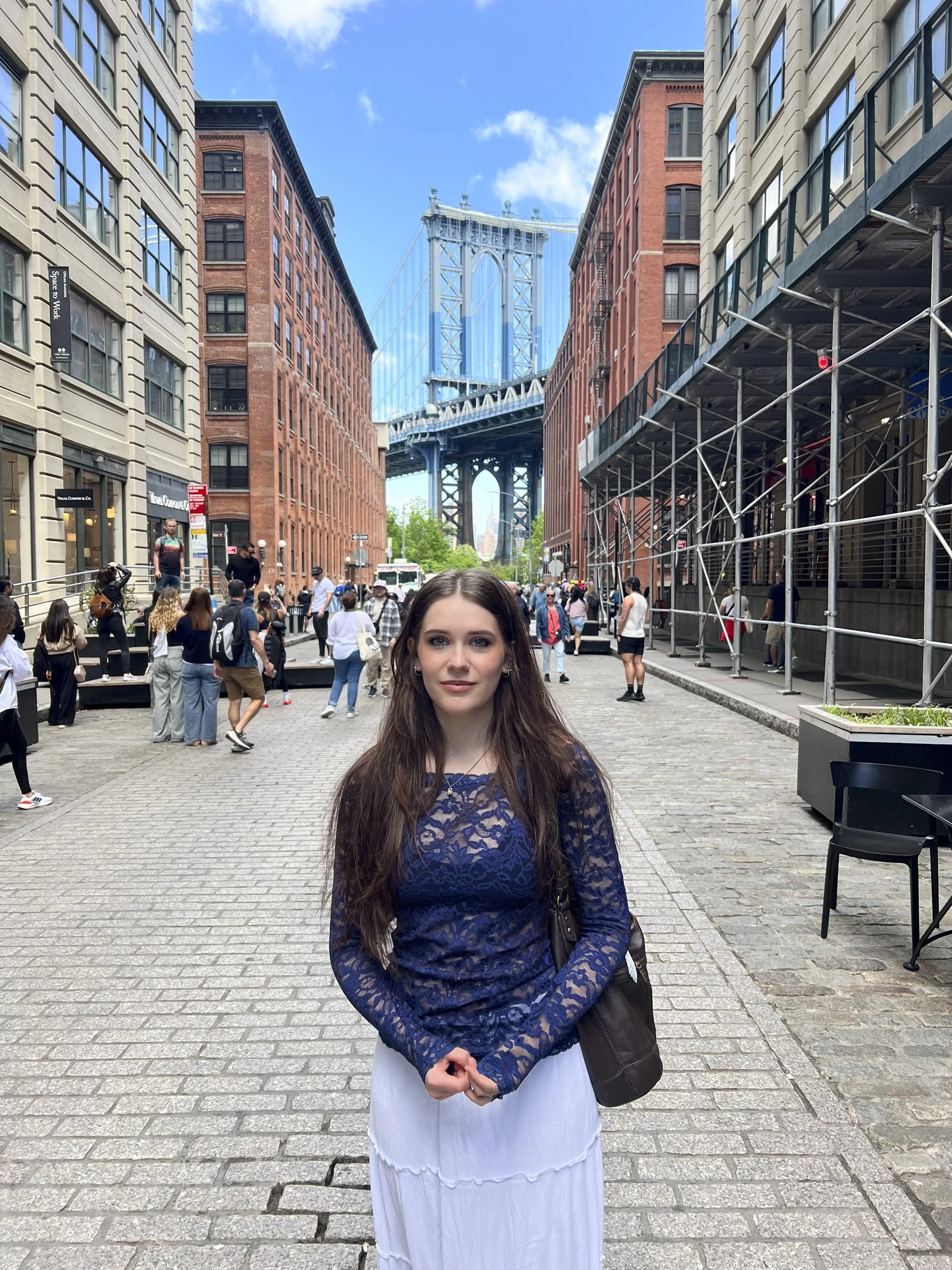 A young woman with long dark hair wearing a blue lace top and white skirt, standing on a cobblestone street in a city with buildings, scaffolding, and the Manhattan Bridge in the background, under a partly cloudy sky.