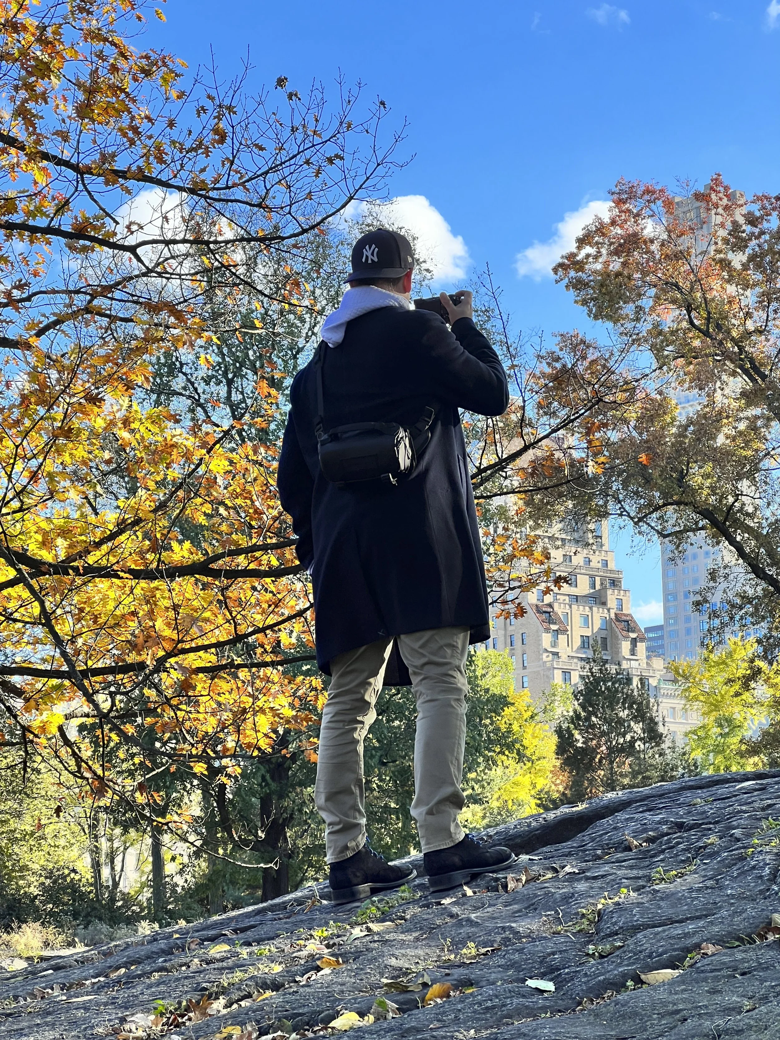 A person (Ken) standing on a rock in Central Park with trees showing autumn colors, taking a photo with a Sony A7cr under a blue sky with some clouds.