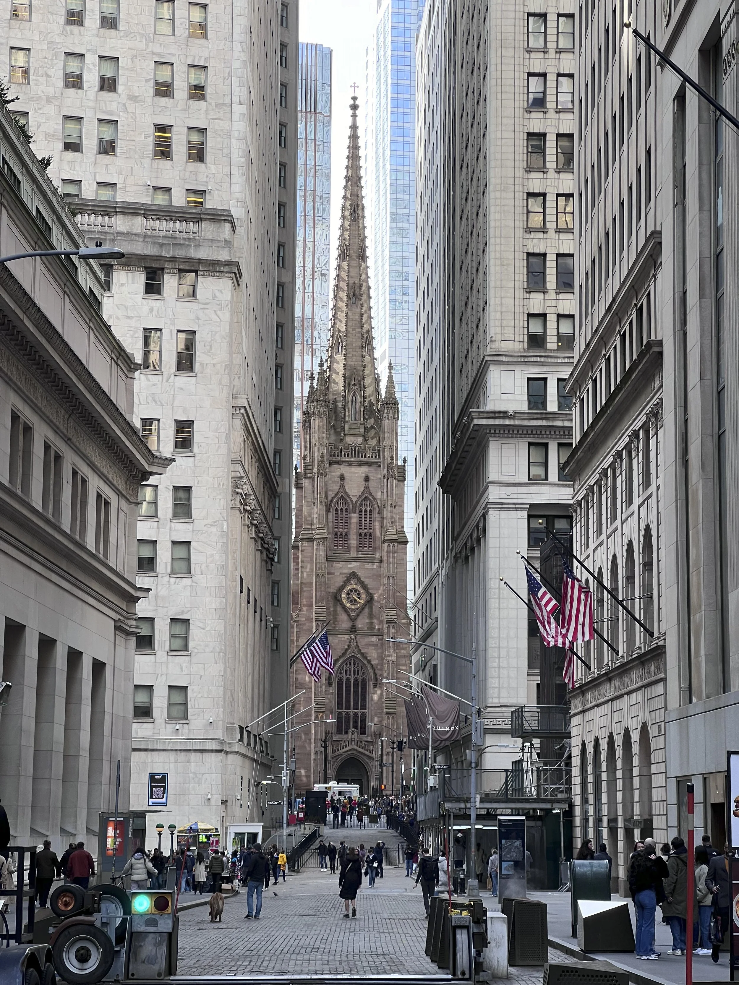 Southern Manhattan street in New York City with the historic church called Trinity centered in the photograph. A tall spire, American flags, and many pedestrians walking.