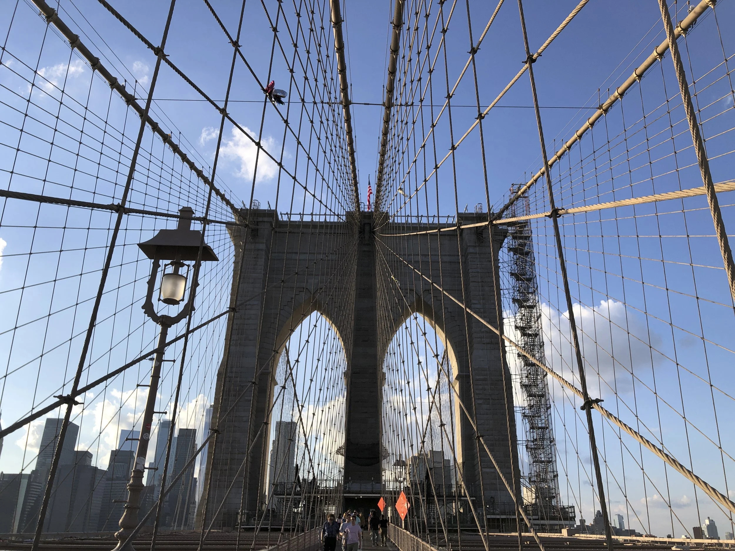 View of the Brooklyn Bridge in New York City showing its stone towers, steel cables, and a blue sky with clouds, a photographic lesson in symmetry and composition.
