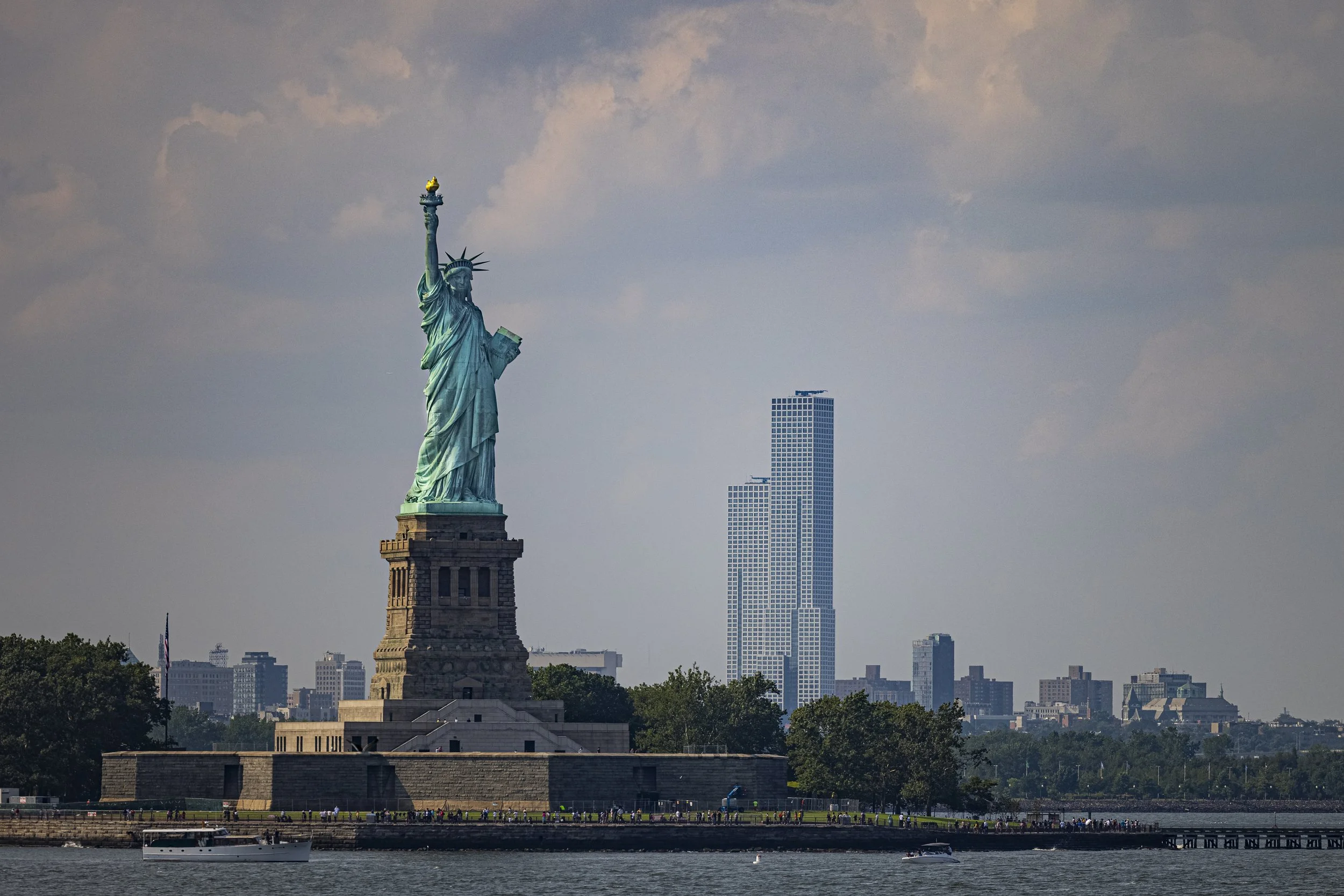 The Statue of Liberty standing on its pedestal, with a cityscape including tall skyscrapers in the background, seen across a body of water with boats.