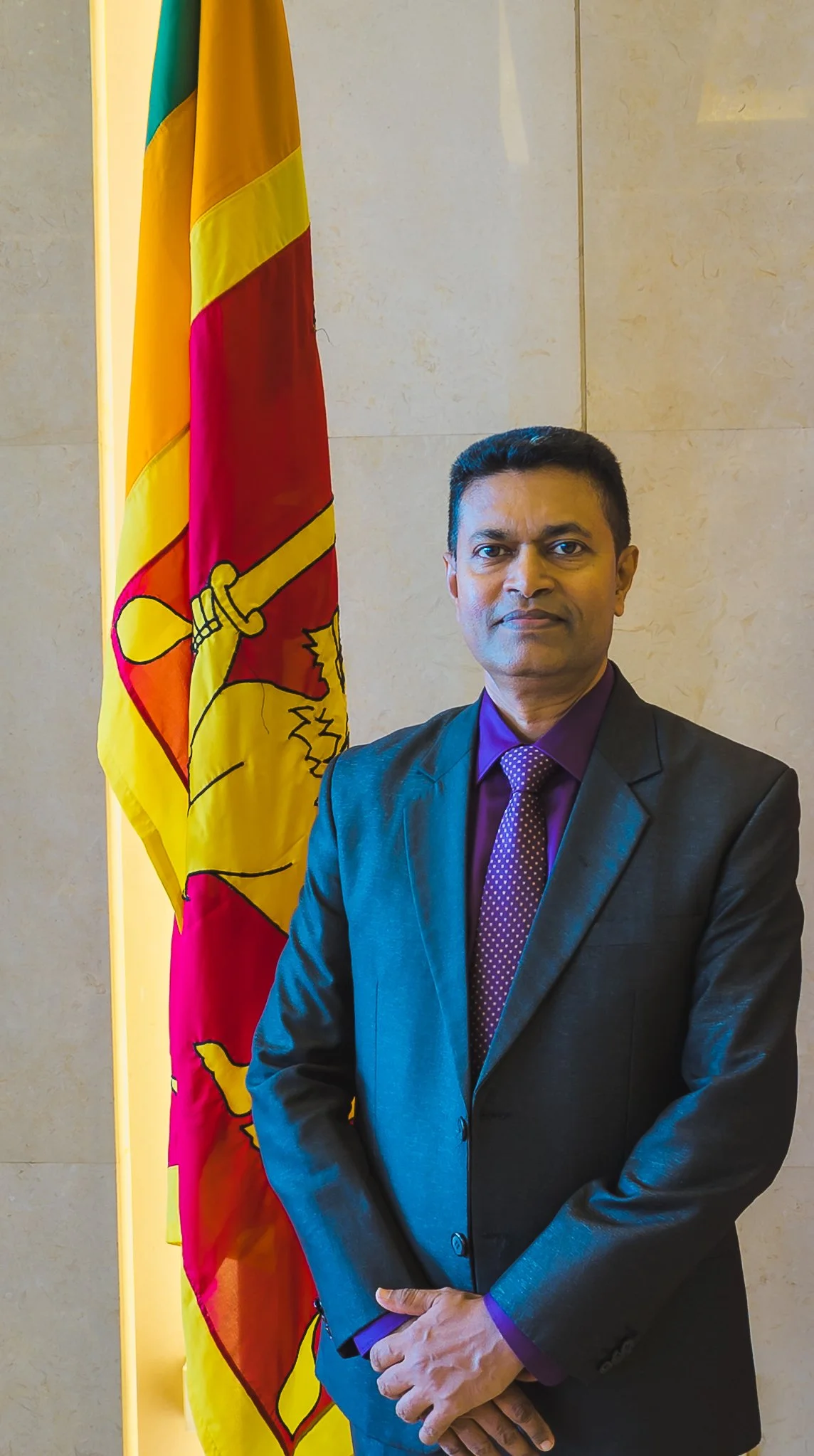 A man in a dark suit with a purple shirt and purple polka dot tie standing in front of a Sri Lankan flag against a beige wall.
