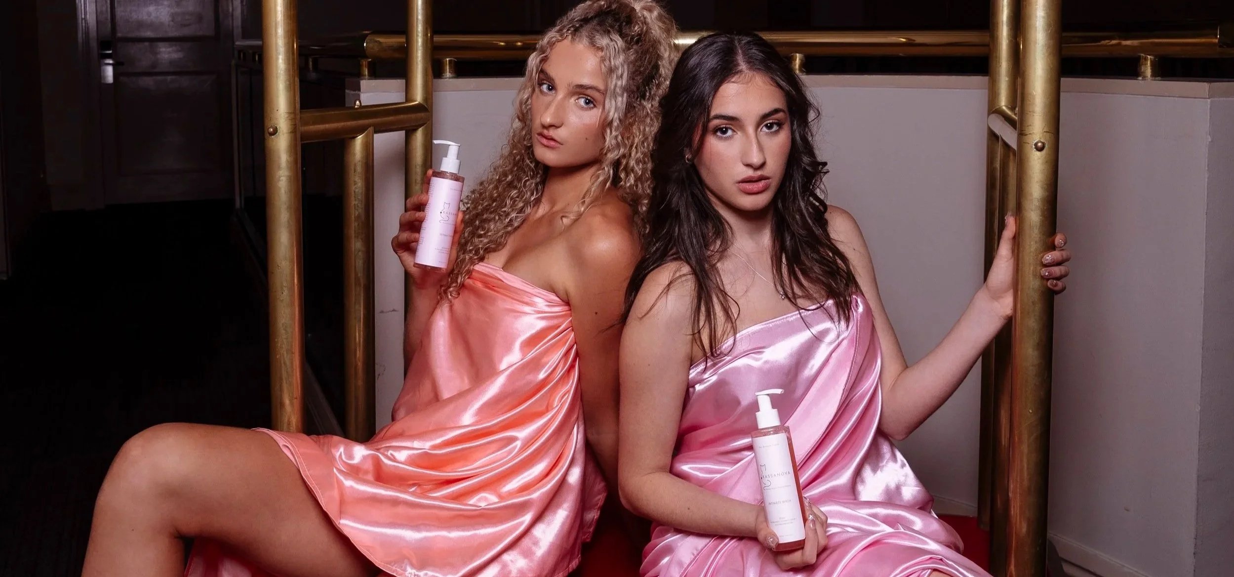 Two women sitting and leaning against a golden metal structure, wearing pink satin outfits, each holding a skincare product with a pump dispenser.