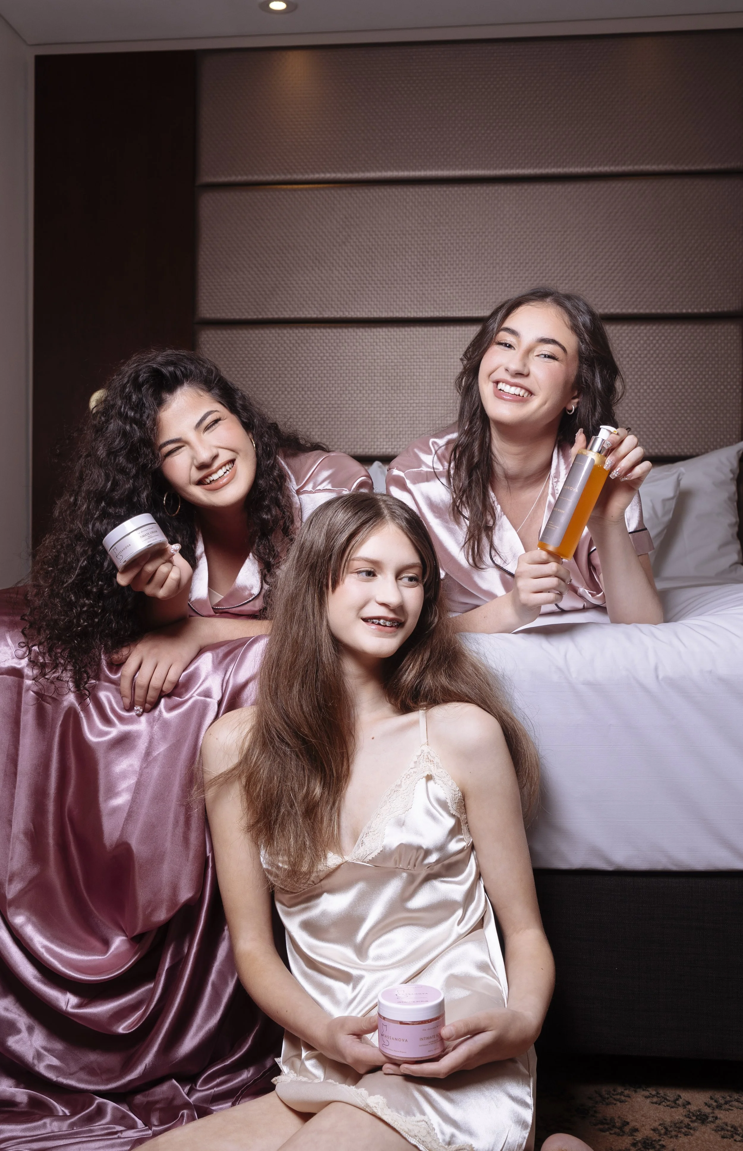 Three women in satin robes enjoying skincare products in a bedroom.