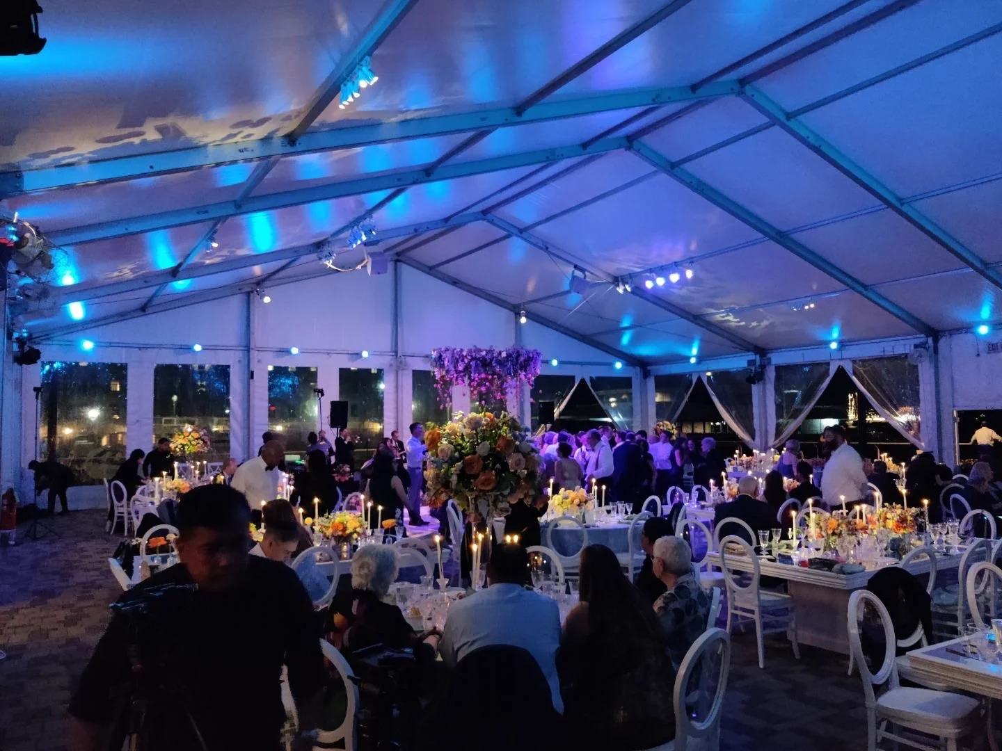 Indoor wedding reception with decorated tables, floral arrangements, and people dancing under a tent with blue lighting.