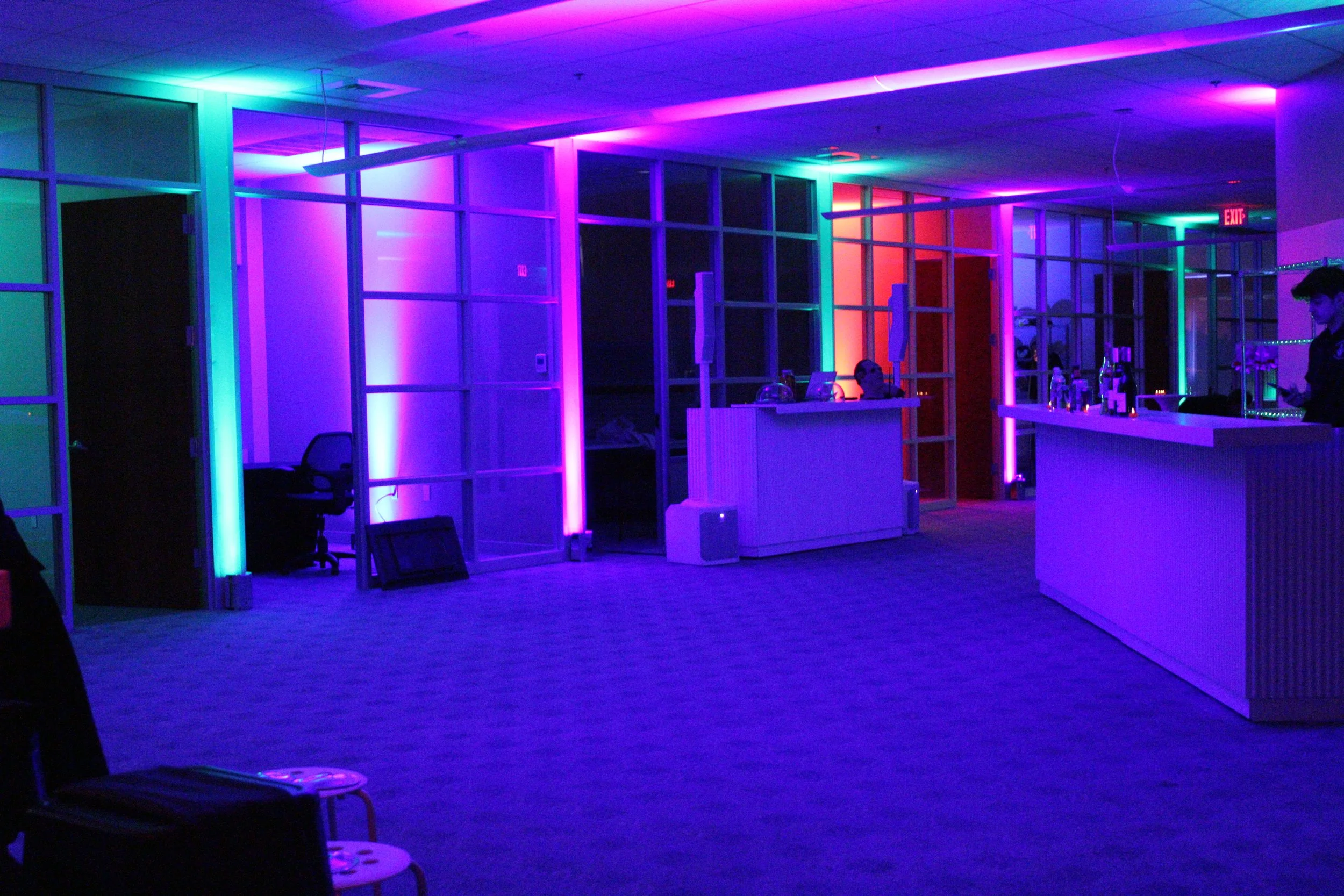 An empty reception area lit with vibrant purple, pink, and blue LED lighting, featuring a white bar counter with bottles, a DJ booth, and glass partition walls.