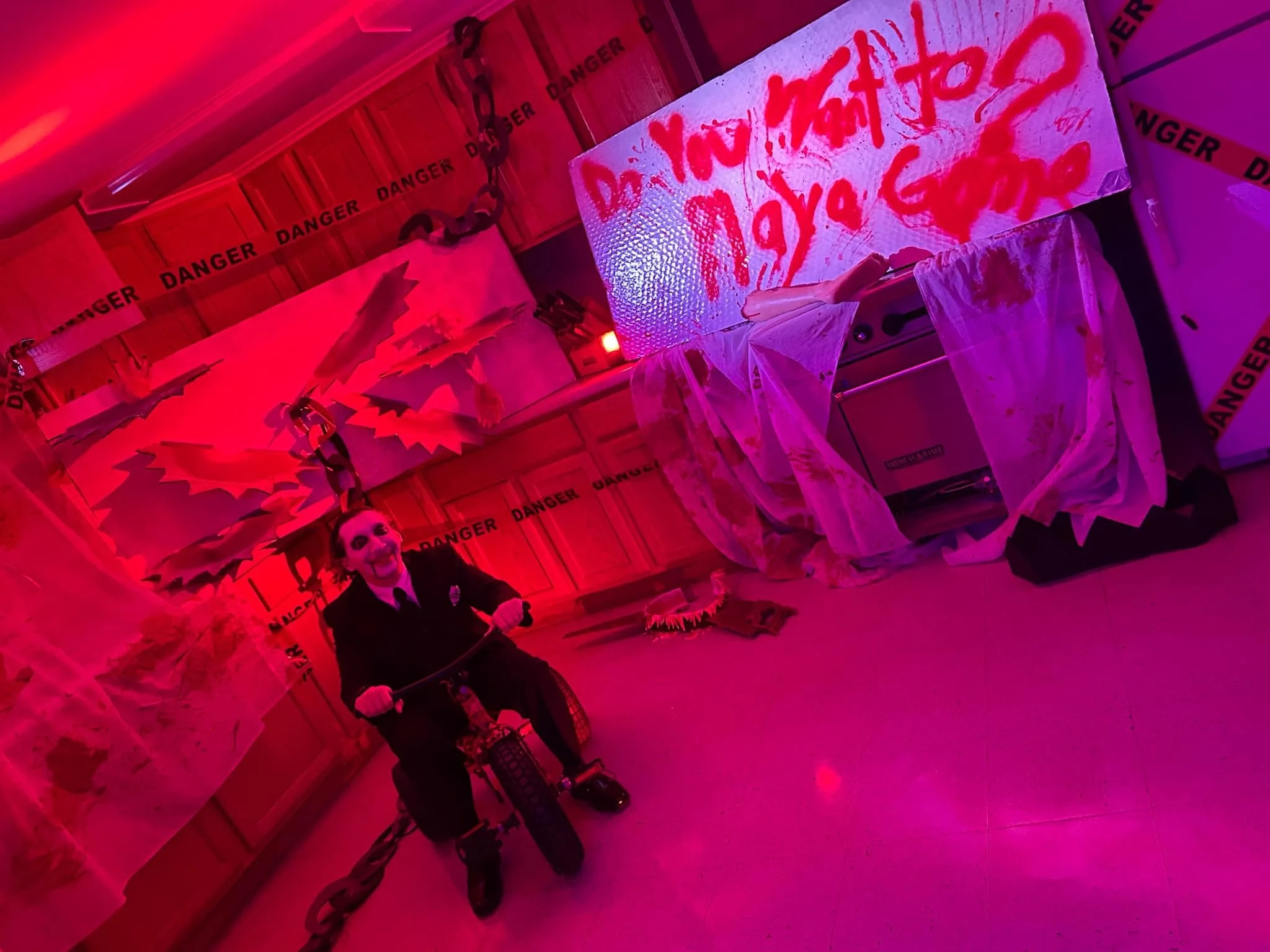 A creepy kitchen scene with red and purple lighting, medical danger tape on the walls, a mannequin with a broad smile riding a tricycle, a large whiteboard with red handwritten messages, bloody cloths scattered on the floor, and various unsettling de