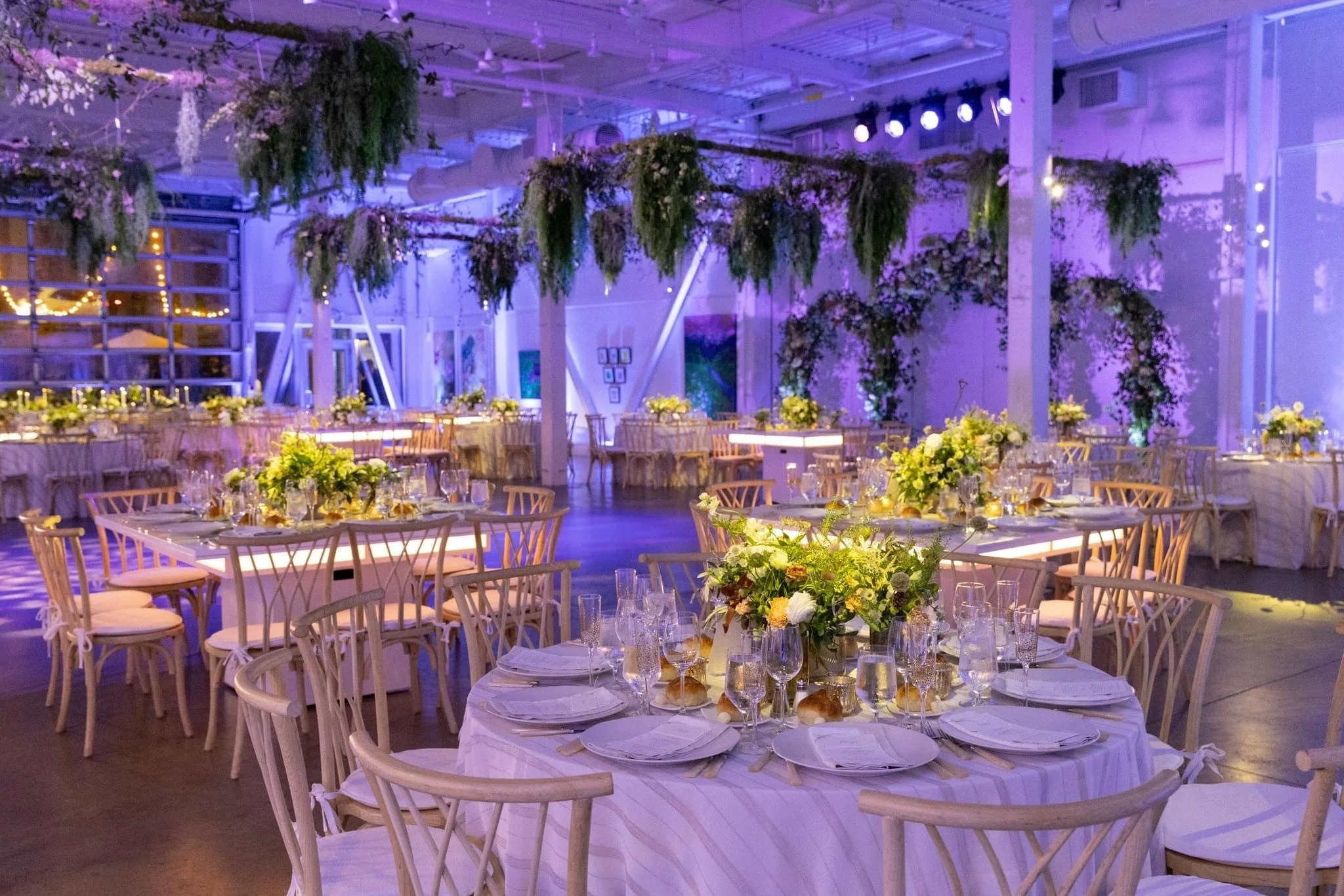 An elegant event space decorated with hanging greenery and fairy lights, featuring round tables with white tablecloths, floral centerpieces, and place settings. The ambiance is soft purple and warm lighting.