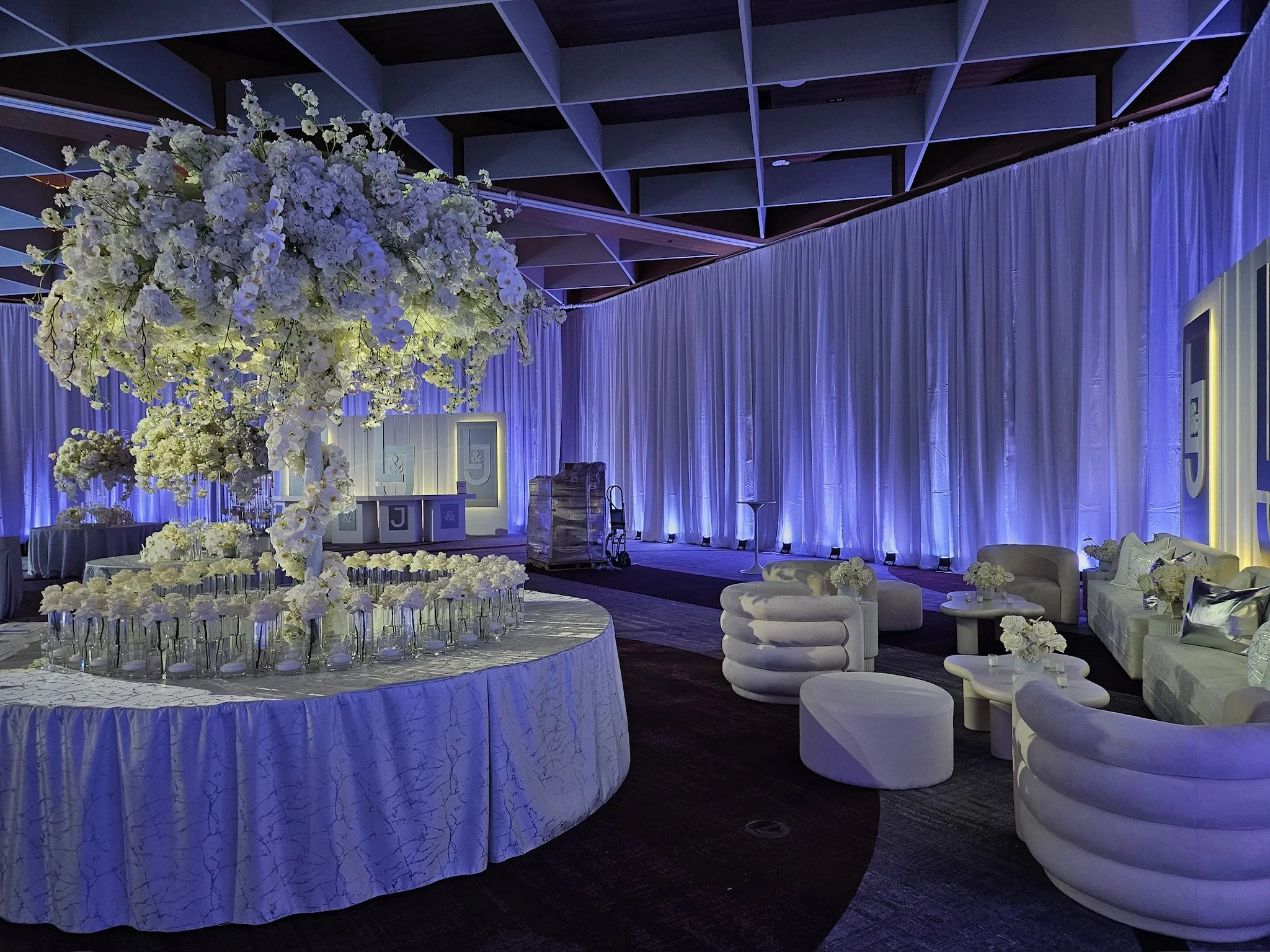 Elegant event space with a large floral centerpiece, white seating, and draped curtains illuminated in purple.