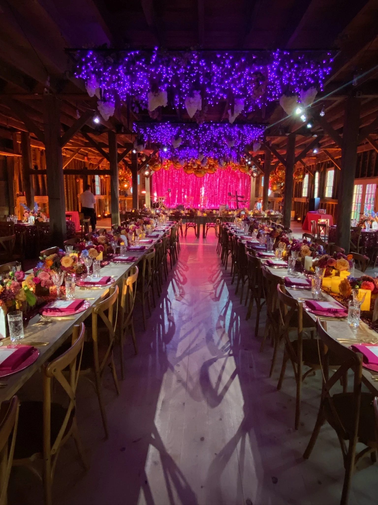 Decorated indoor wedding reception with long banquet tables, floral centerpieces, pink napkins, and purple and yellow lighting effects. Purple string lights hang from the ceiling with floral arrangements, and a pink curtain backdrop on stage.