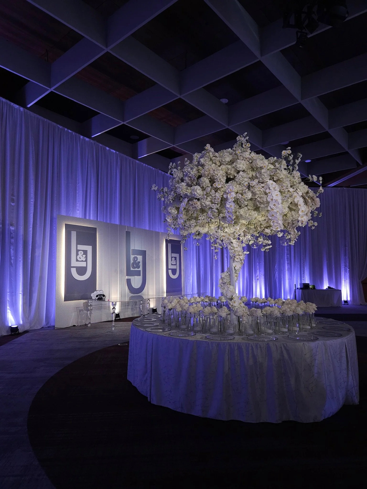 Elegant event setup with a large floral centerpiece on a round table, surrounded by glasses, in front of a backdrop with J & logo, lit with purple lighting.