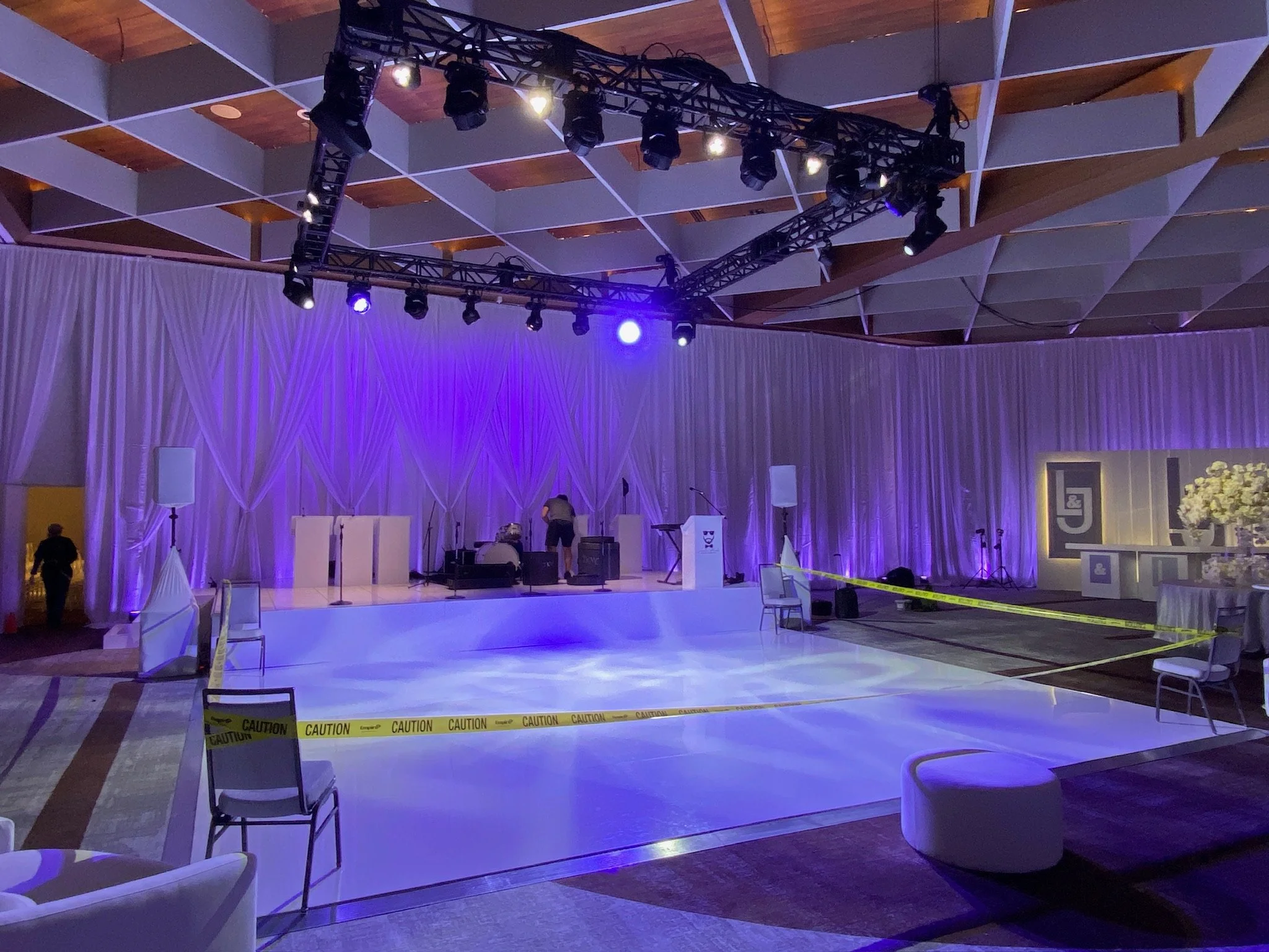 Event setup stage with purple lighting, white draped backdrop, sound and lighting equipment, caution tape on the floor, chairs, and tables in a large indoor event space.