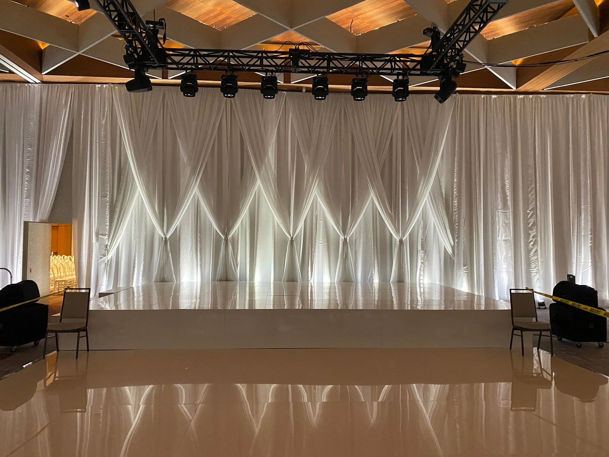 Empty stage with white drapes and curtain backdrop, lighting truss overhead, chairs and suitcases on either side, and a polished floor reflecting the background.