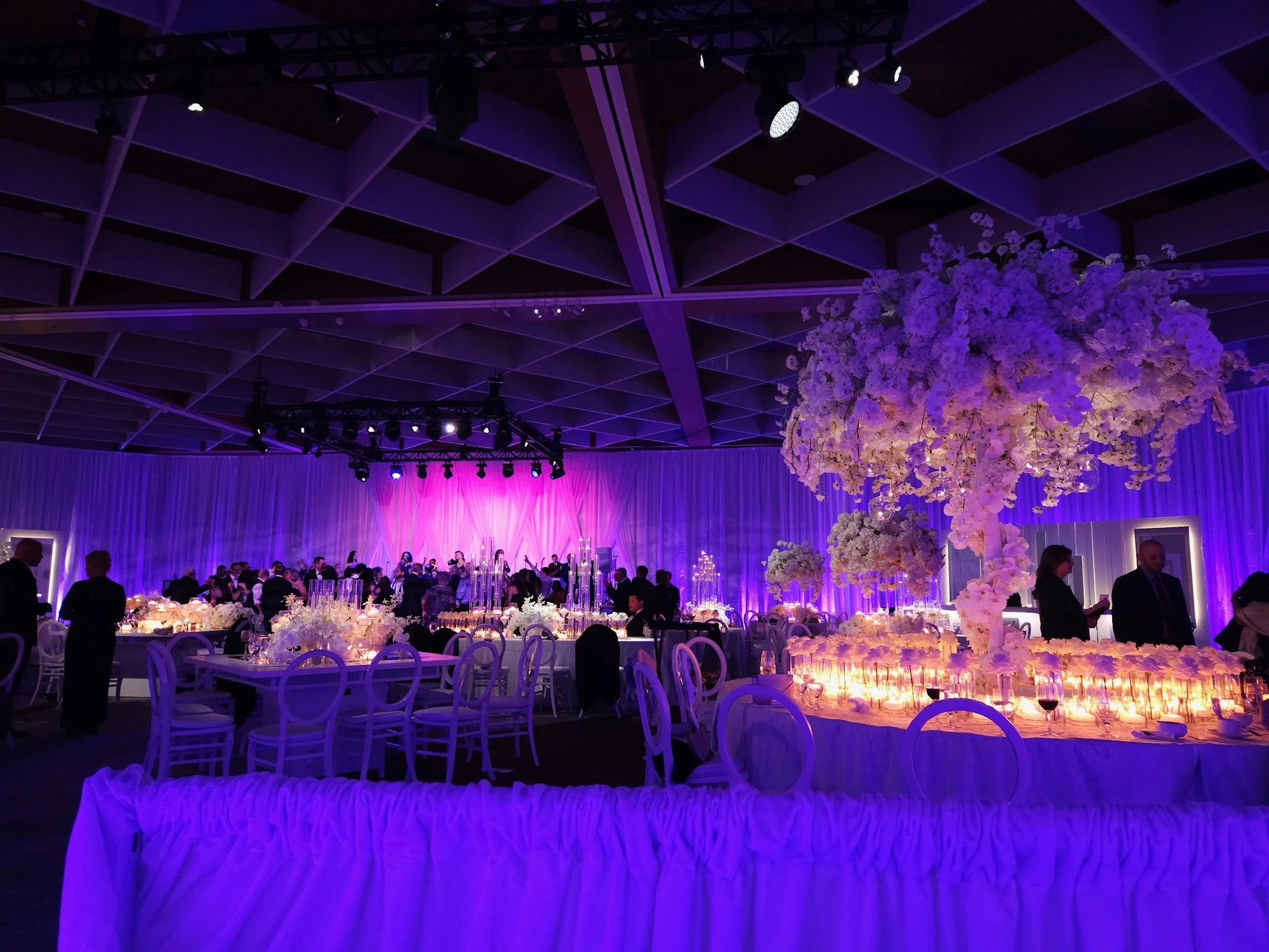 Elegant event hall decorated with purple lighting, large floral centerpieces, and dining tables with candles, set for a celebration or banquet.