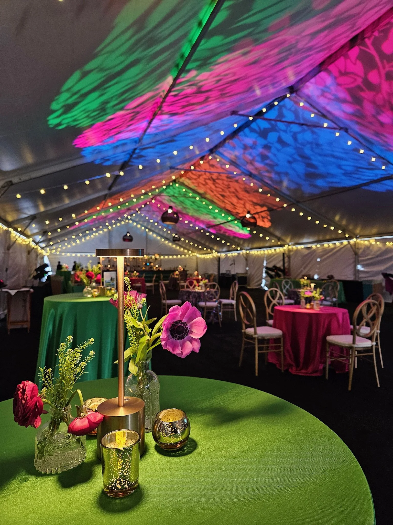 A decorated event tent with colorful ceiling lighting, string lights, and tables with pink and green tablecloths, floral centerpieces, and candle holders.