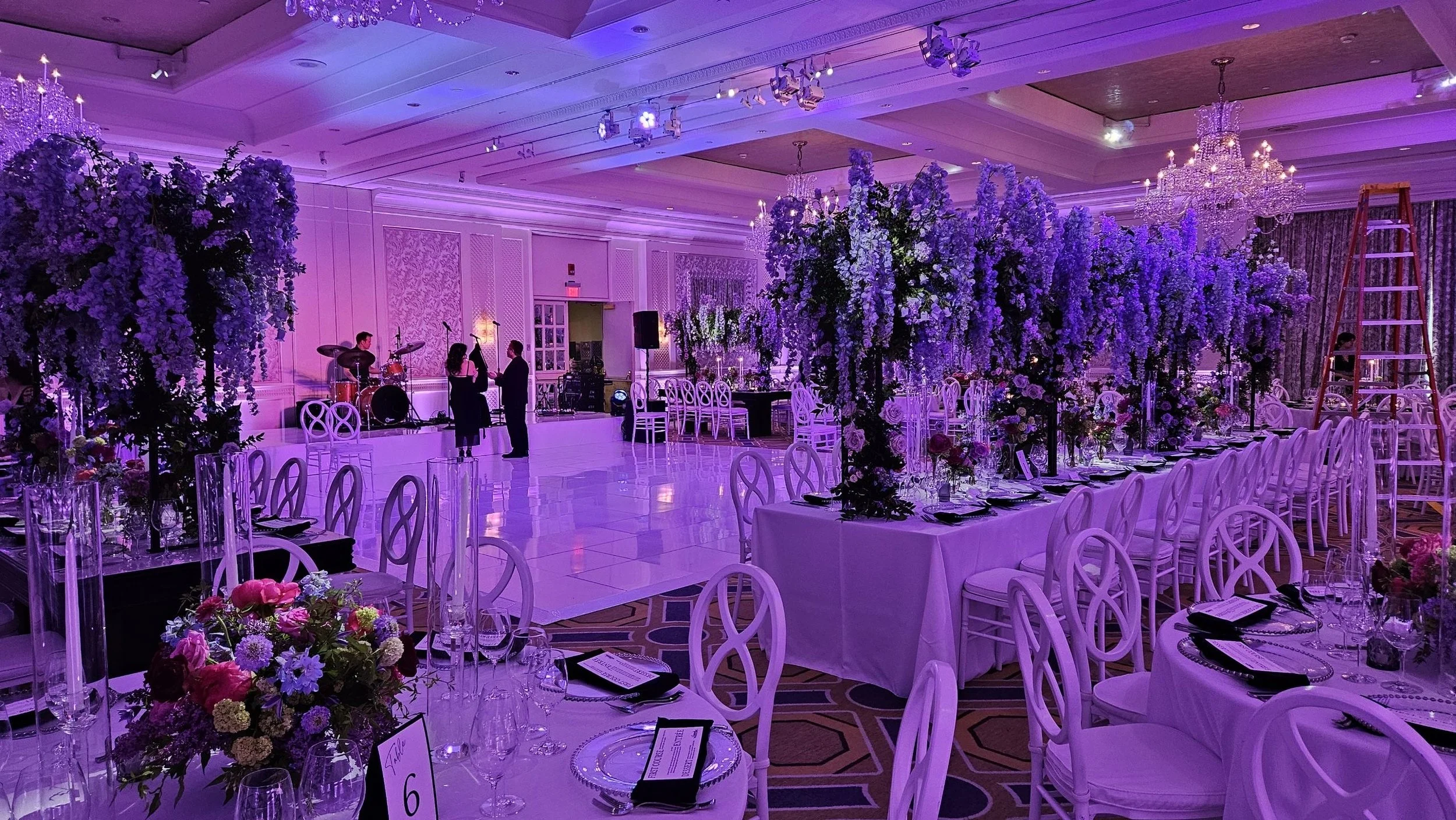 Elegant banquet hall decorated with purple and white flowers and crystal chandeliers, set for a formal event with round tables, chairs, and a dance floor, with a live band playing in the background.