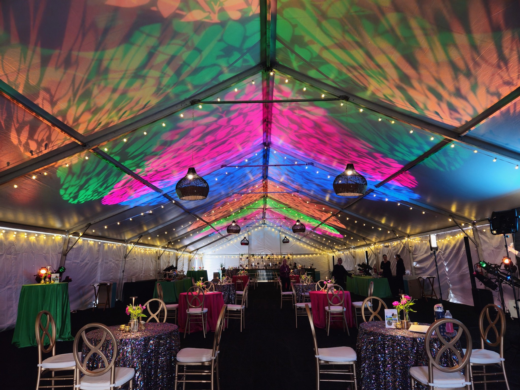 Decorated event tent with multicolored lighting on ceiling, string lights, and round tables with sequin tablecloths and floral centerpieces.