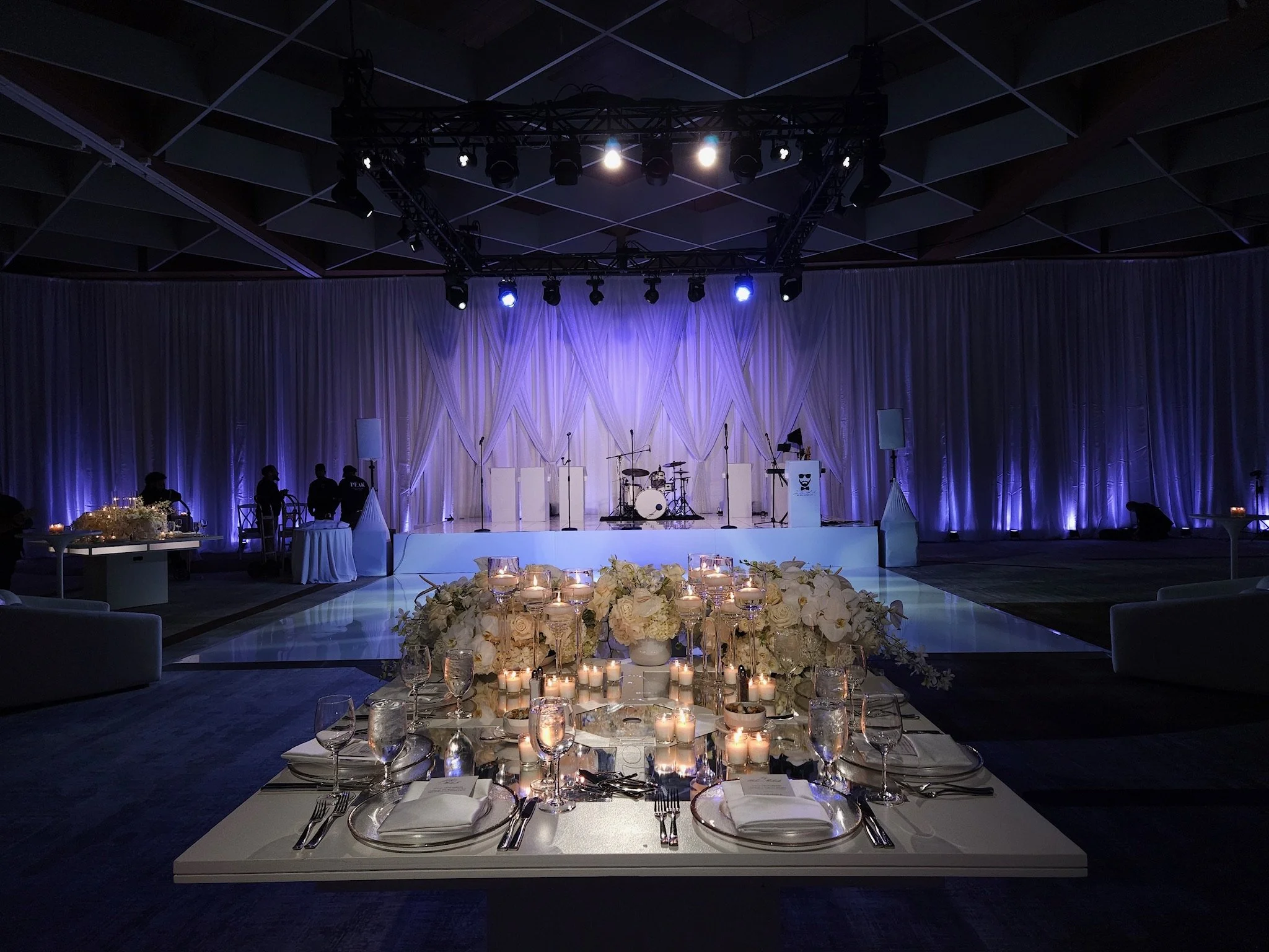 Elegant event space with a stage, musical instruments, and a dining area decorated with white flowers and candles, illuminated with soft purple lighting.