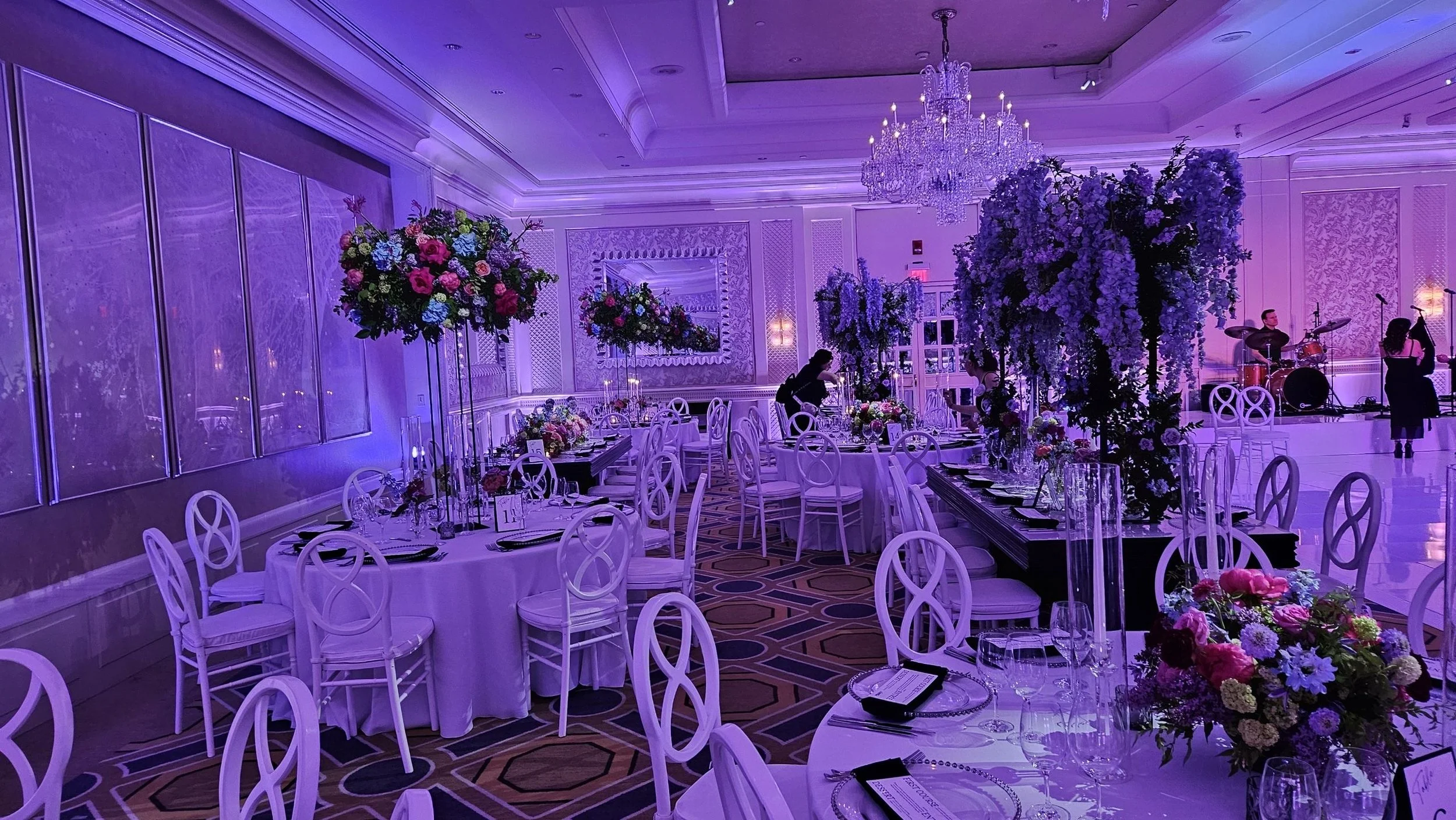 Elegant event hall decorated with purple lighting, large floral centerpieces, and round tables covered with white tablecloths, set for a formal occasion with glassware and place settings. Musicians are setting up on stage in the background.