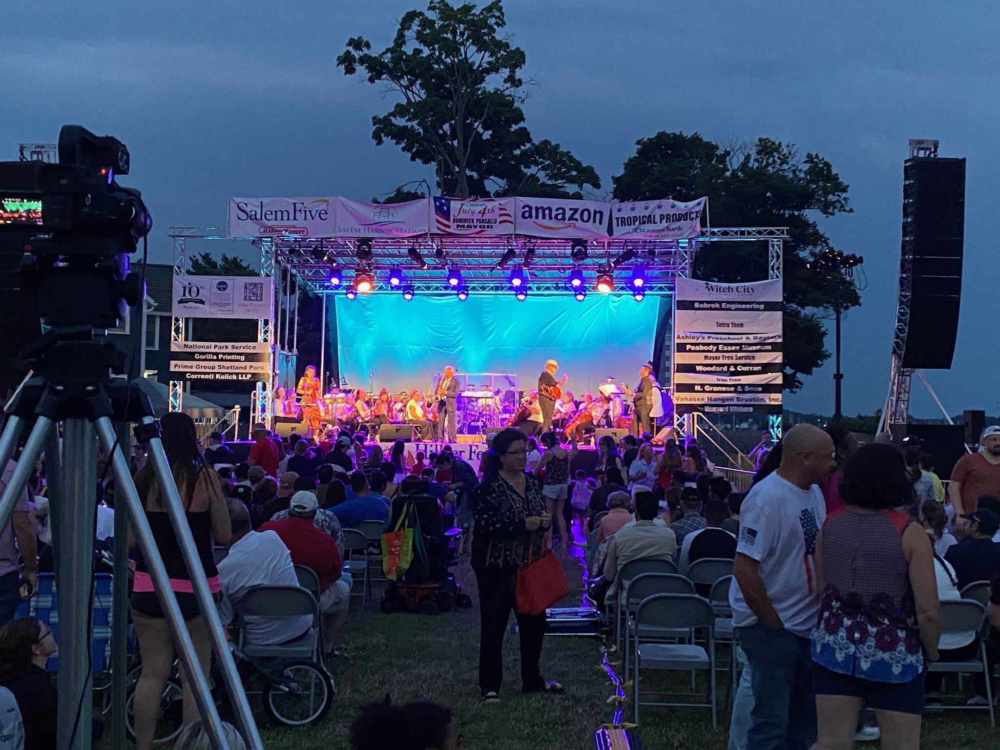 Outdoor concert at dusk with a stage illuminated in blue lights, a live band performing, and an audience seated and standing in front of the stage.