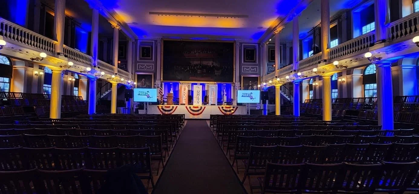 Empty auditorium with rows of chairs facing a stage decorated with patriotic banners, two large screens displaying 'Project' and 'Quality', and an American flag in the center.