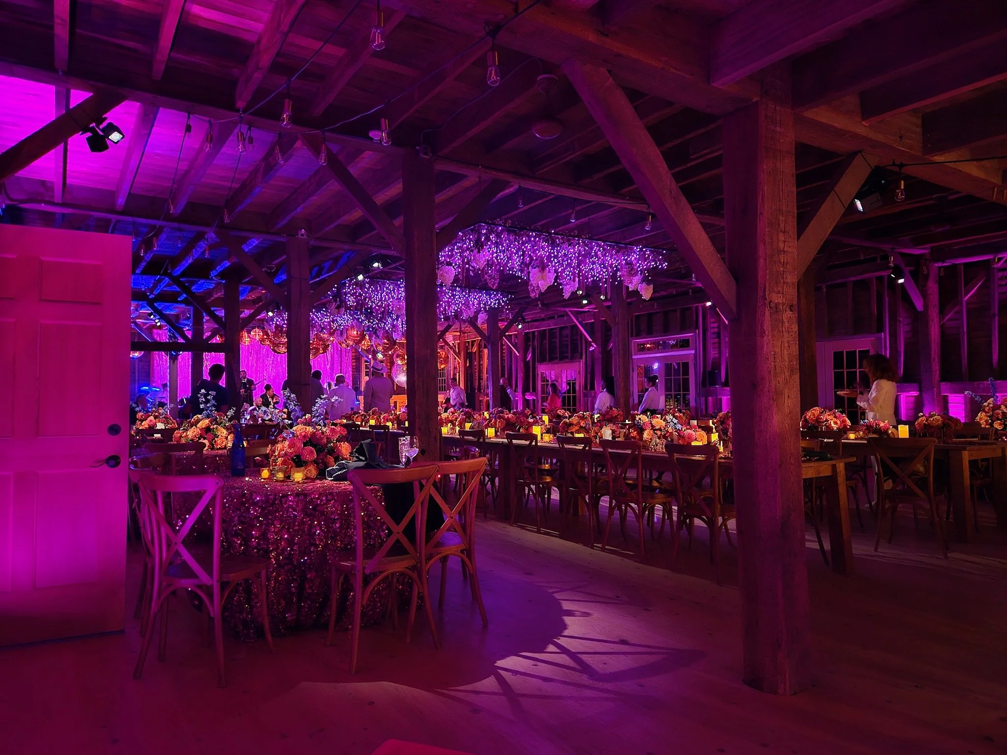 Interior of a wedding reception hall with purple and pink lighting, decorated tables with floral centerpieces, and guests mingling under hanging string lights.