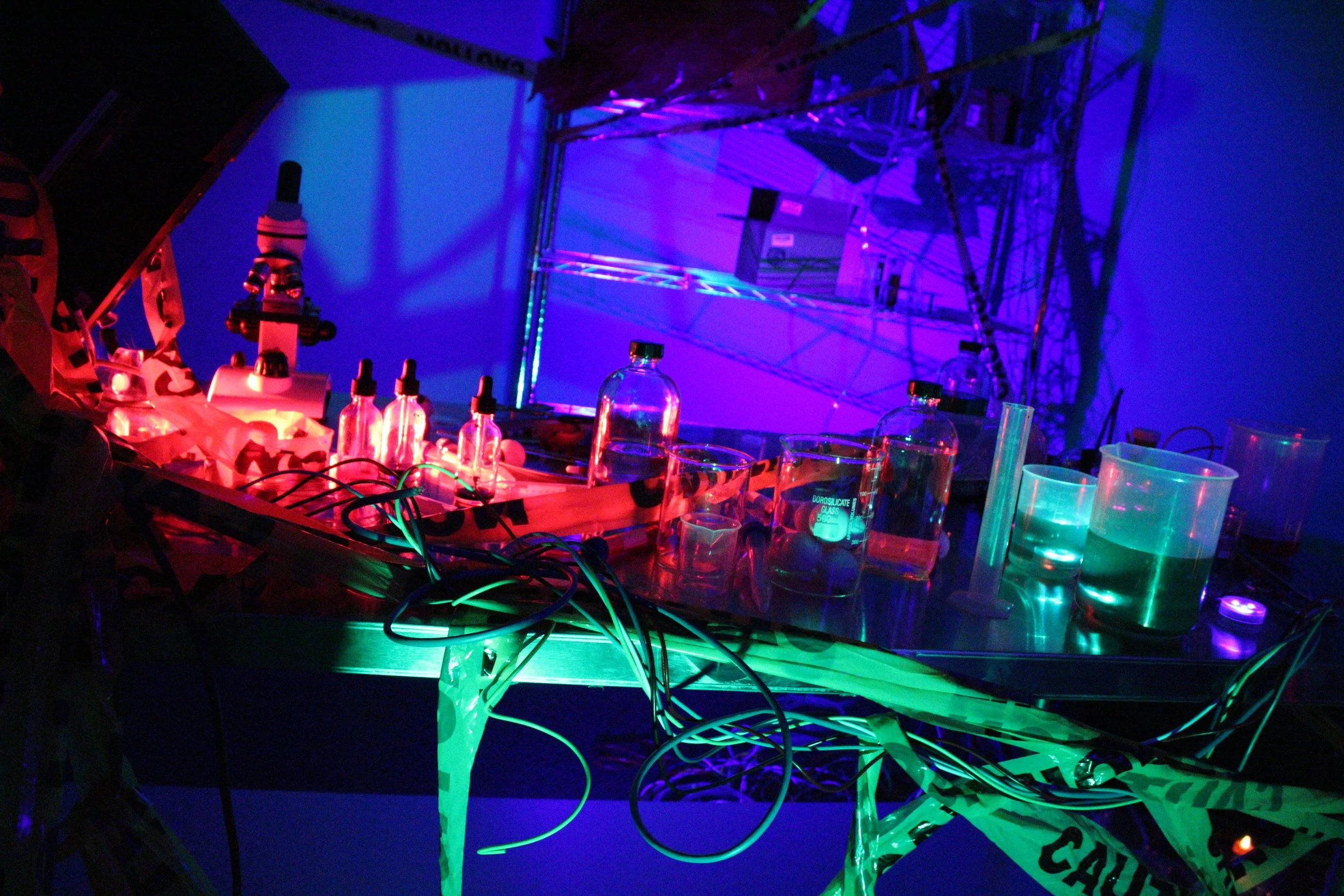 A science lab setup with fluorescent liquids, bottles with droppers, and electronic equipment illuminated by neon lights in RGB colors.