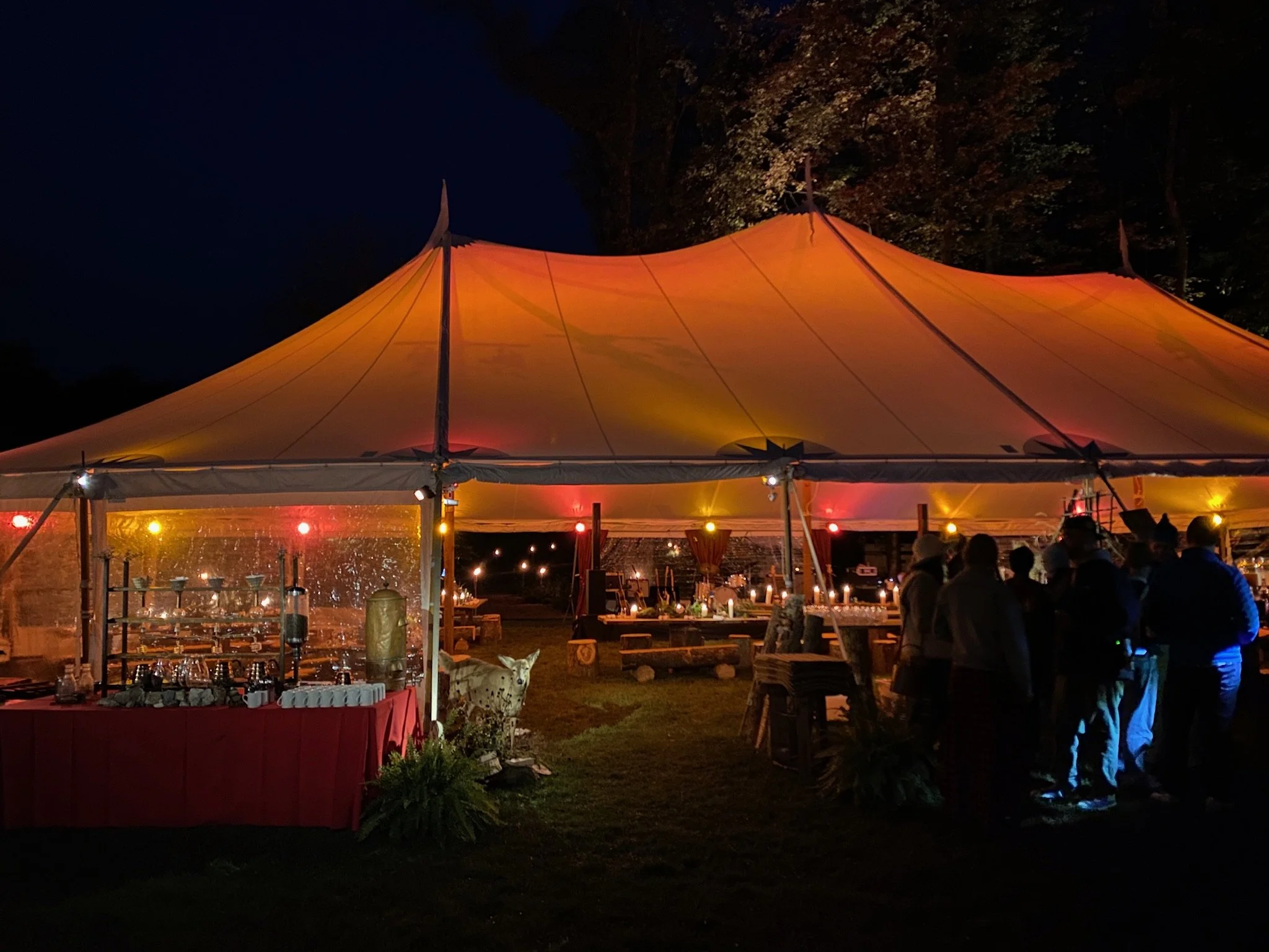 Nighttime outdoor event under a large yellow and orange striped tent with people gathered around in a line, string lights, and food stations.