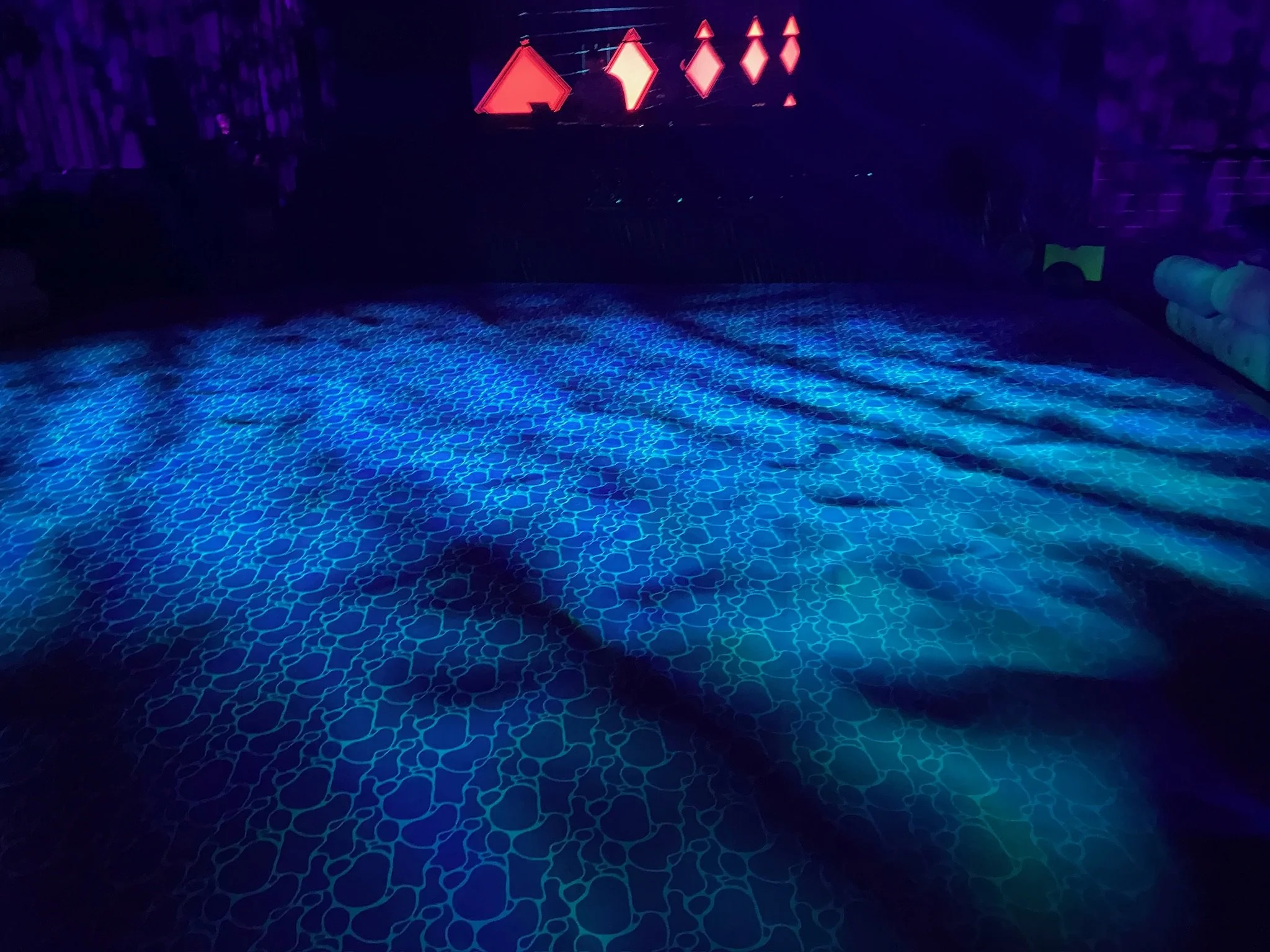 Indoor scene with blue patterned floor and neon red signs on the wall in the background, possibly a nightclub or entertainment venue.