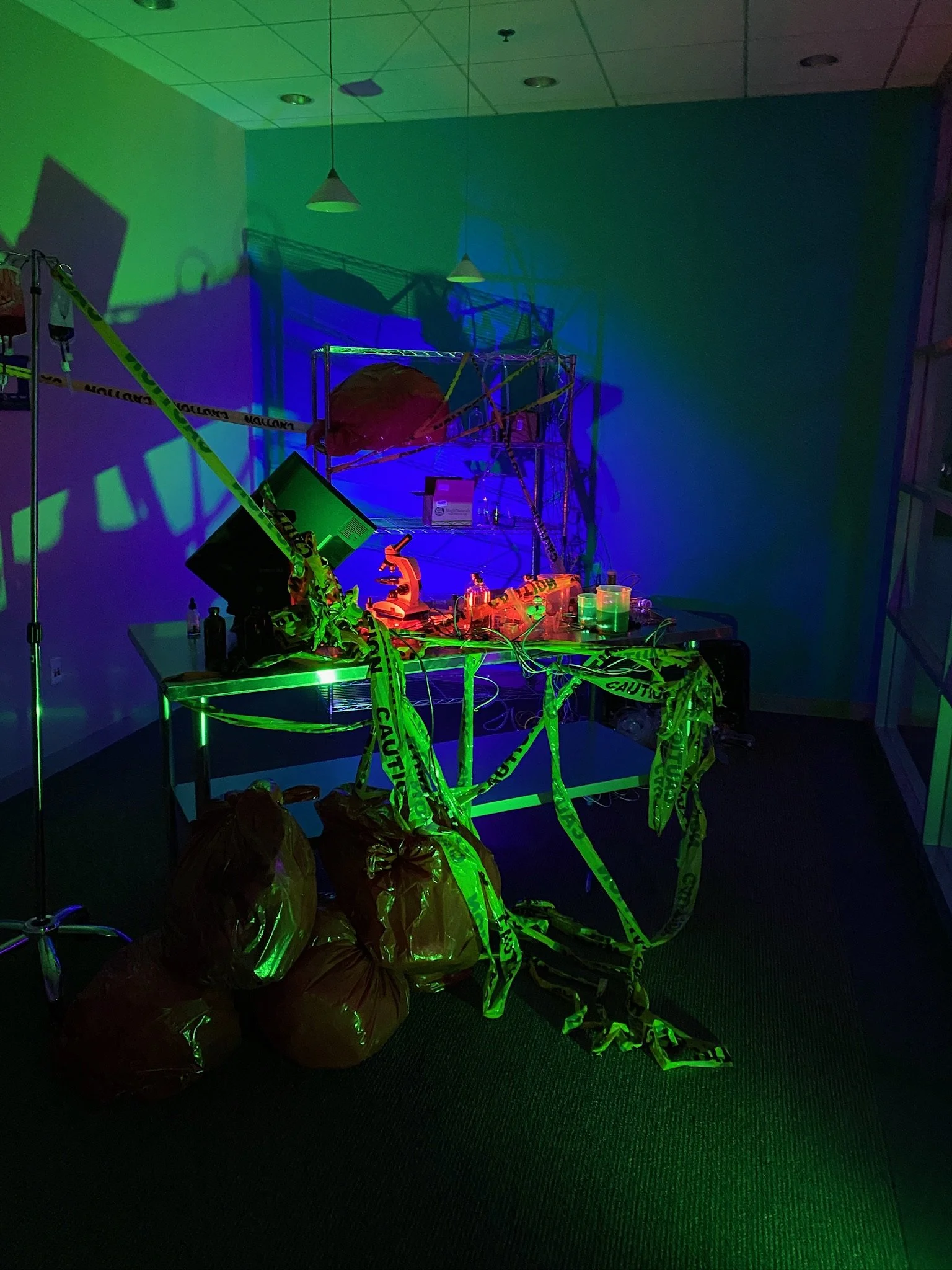 A room with a table and shelves covered with caution tape, green and blue lighting, and scattered items including bags, with a science or investigation setup.