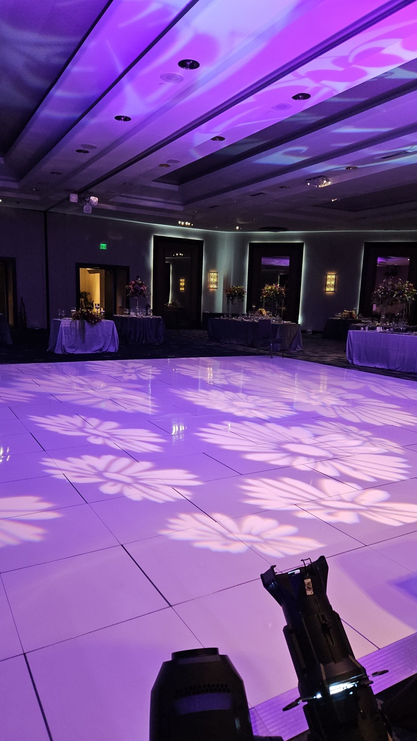 Empty event hall with purple floral patterns projected onto the dance floor, decorated tables with flower arrangements, and dim lighting.