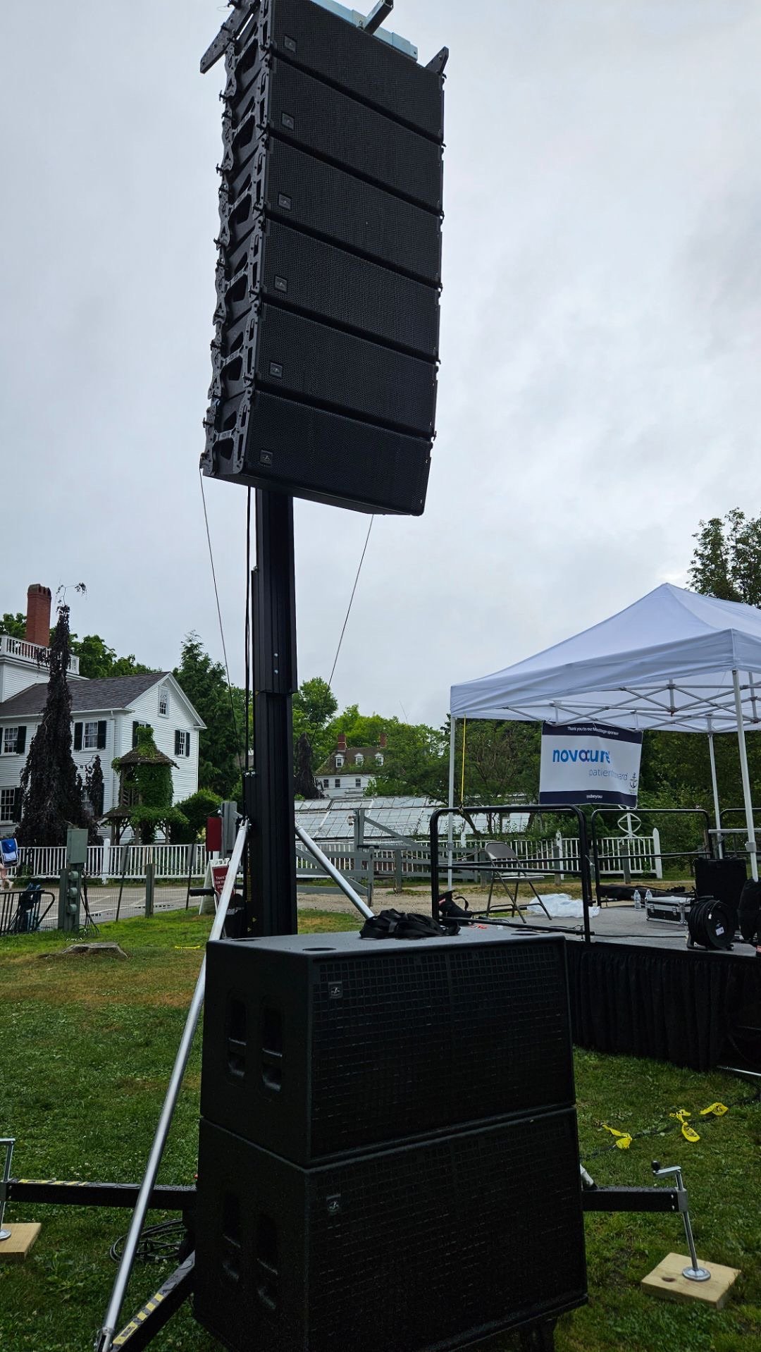 A large outdoor speaker system setup with two stacked speaker units on a stand, on a grassy area near a stage with a white canopy tent and chairs, with residential houses in the background.