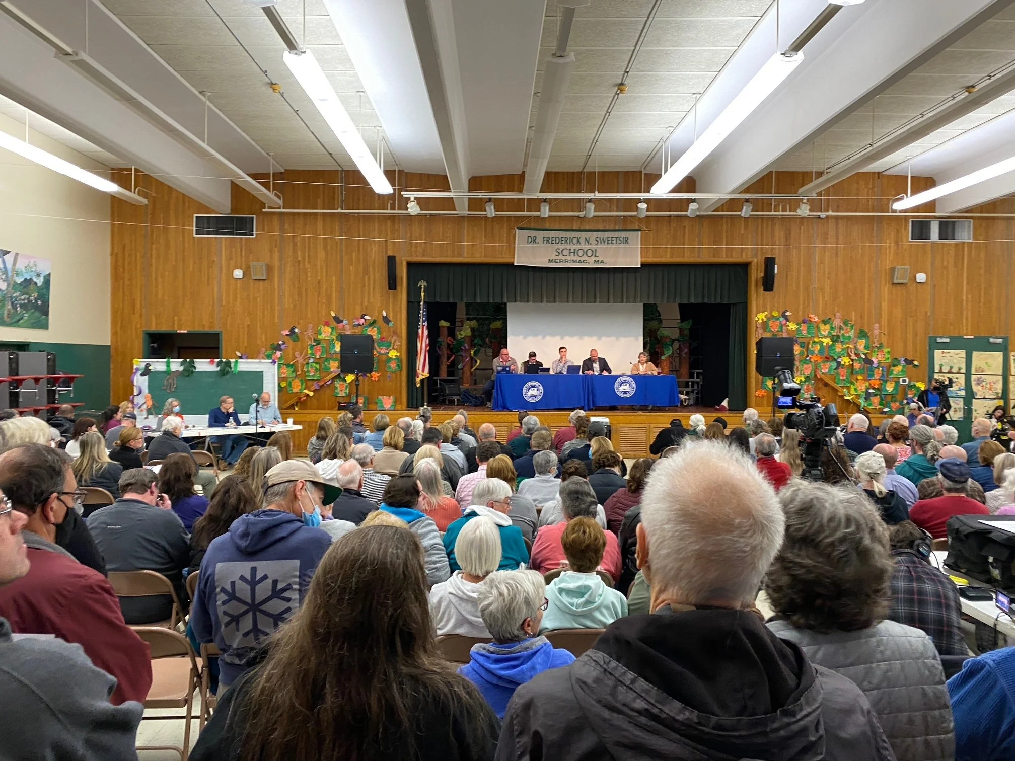 A large gathering of people seated in an auditorium listening to a panel of speakers at a school event. The school stage is decorated with colorful artwork, and a banner overhead reads 'Dr. Frederick N. Sweetser School, Merrimac, MA.'