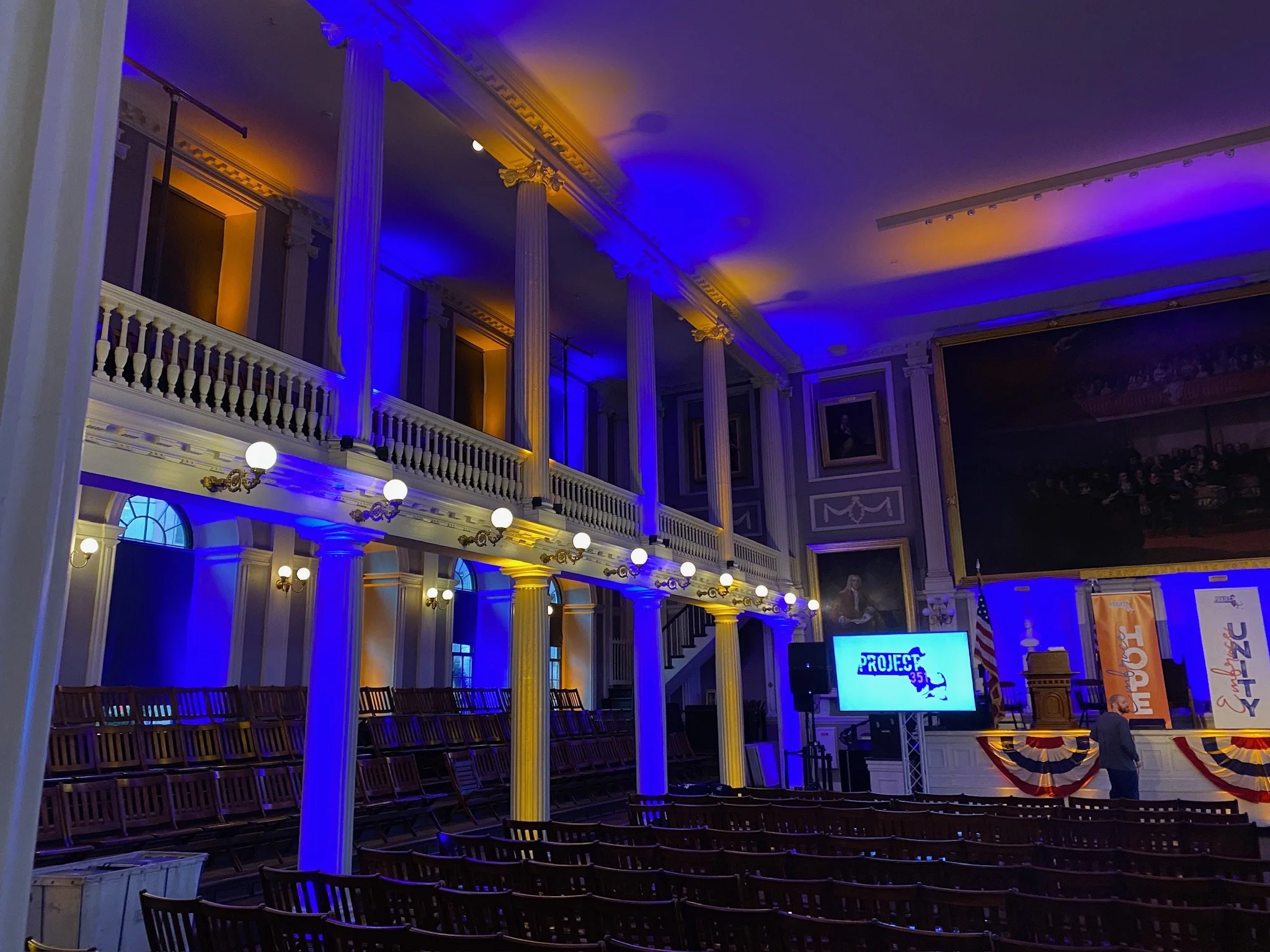 Interior of a grand auditorium or concert hall with ornate columns and balconies, decorated with yellow and blue lighting, and a stage with banners and a large screen displaying 'PROJECT 35.'