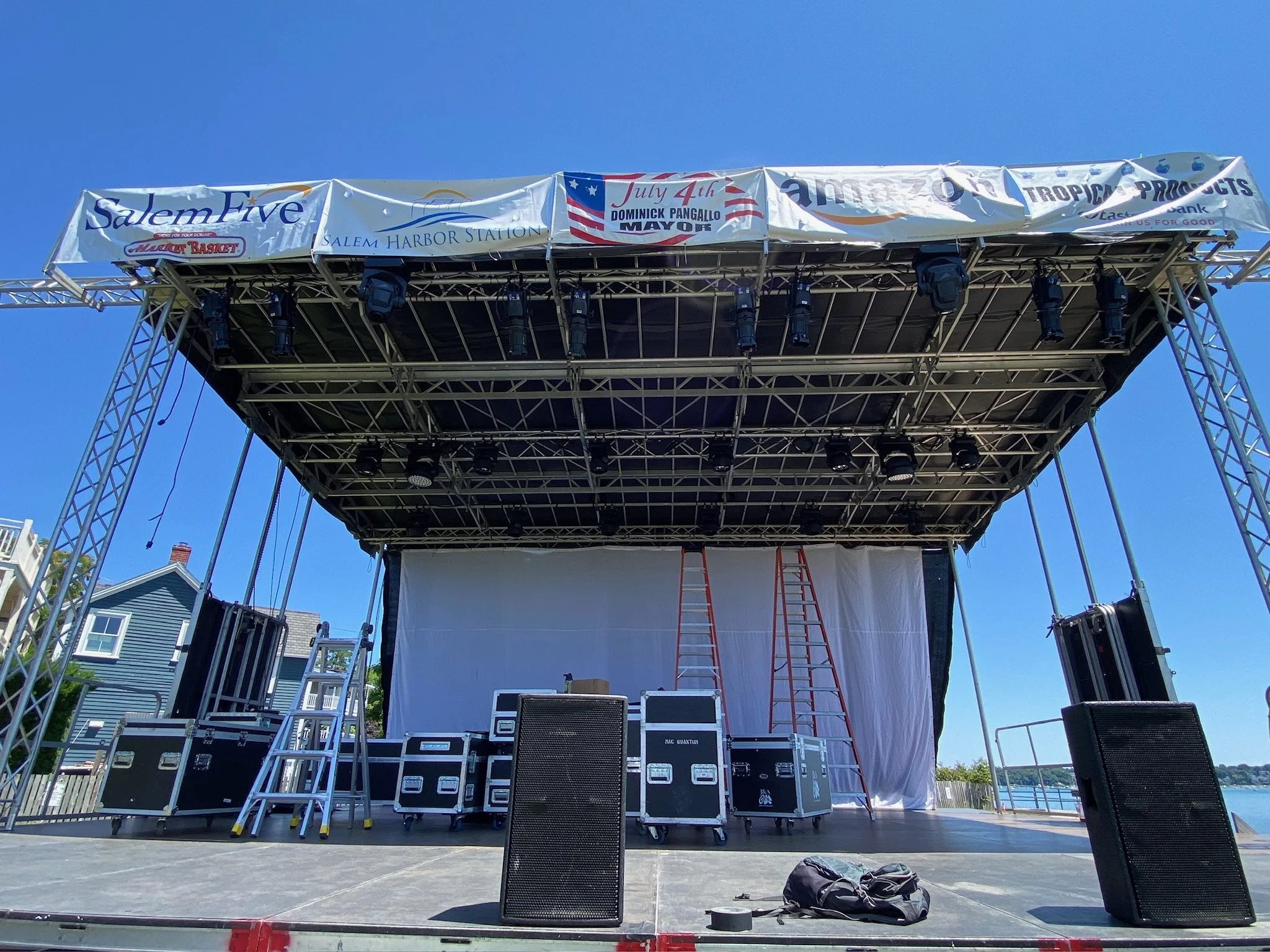 Empty outdoor stage with black equipment cases, ladders, and a white backdrop, set up for a performance with banners above advertising local sponsors and a July 4th event.