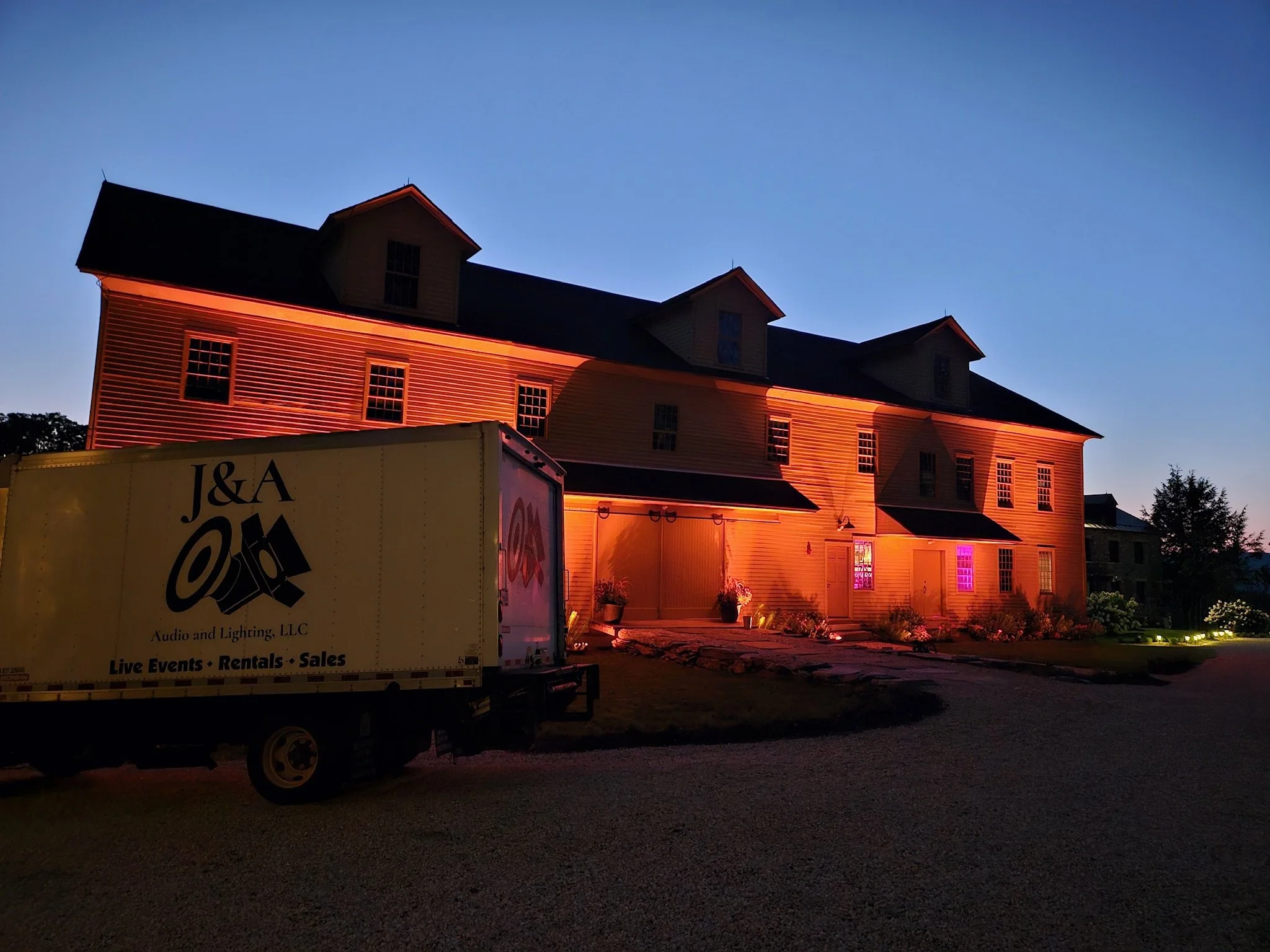 A large three-story house with horizontal siding illuminated with pink and orange lighting at dusk. There are three small dormer windows on the roof and two illuminated windows on the second floor. In front, a truck labeled 'J&A Audio and Lighting, L