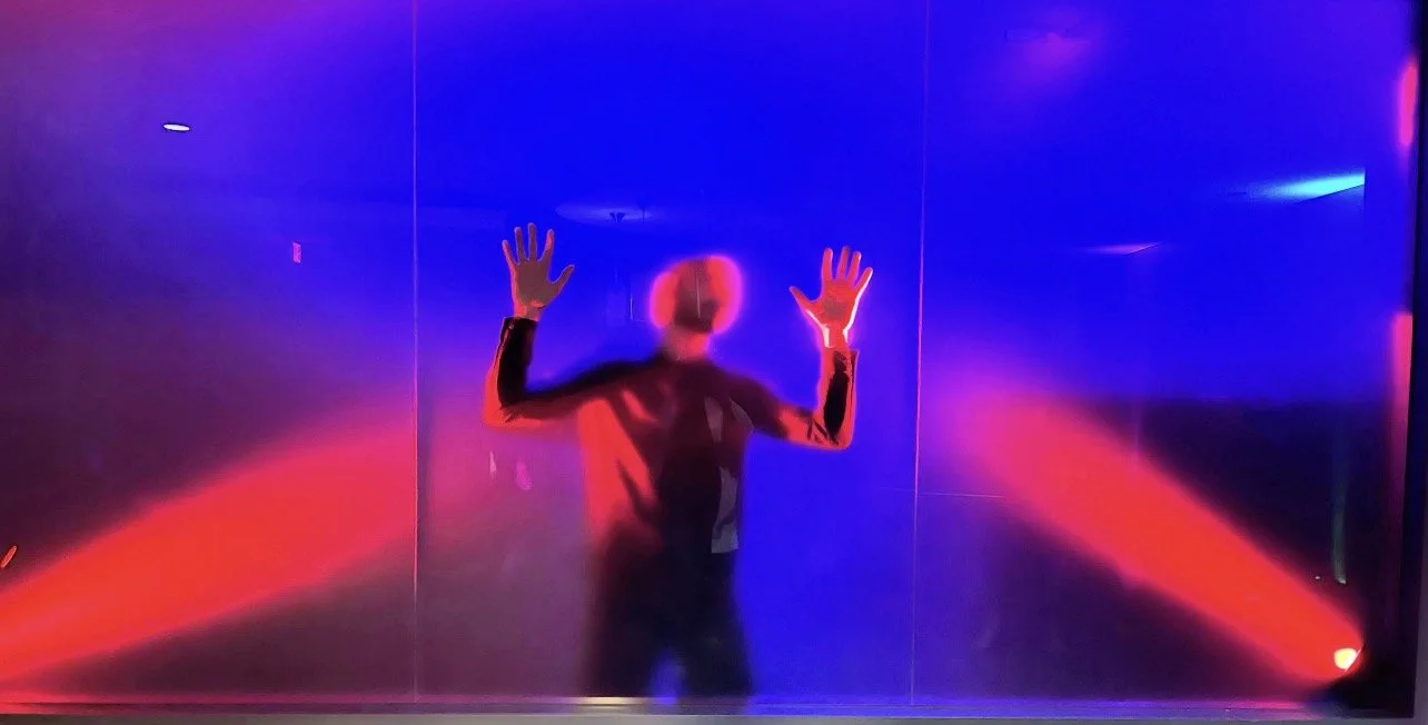 Person standing behind a glass wall illuminated with red and blue lights, with their hands raised and pressed against the glass.