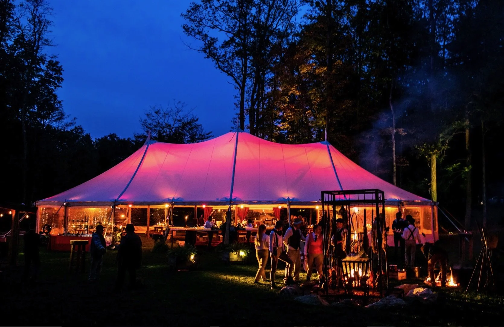 A large pink and purple event tent illuminated at night in a wooded outdoor setting, with people gathered around outside and inside.