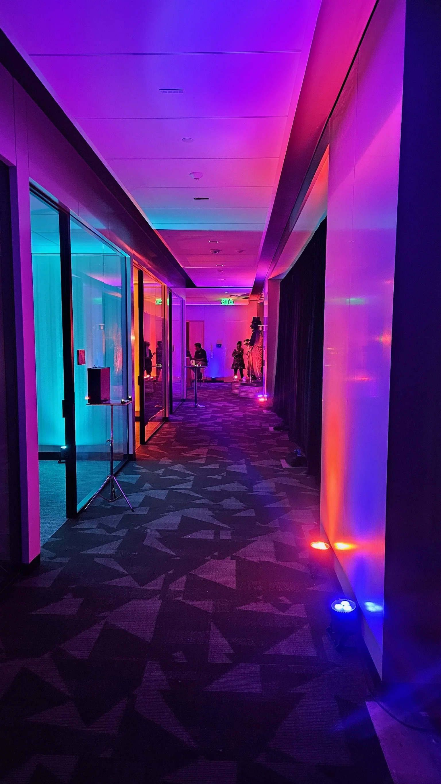 Colorful indoor hallway with purple, blue, and red lighting, glass doors, and people in the distance.