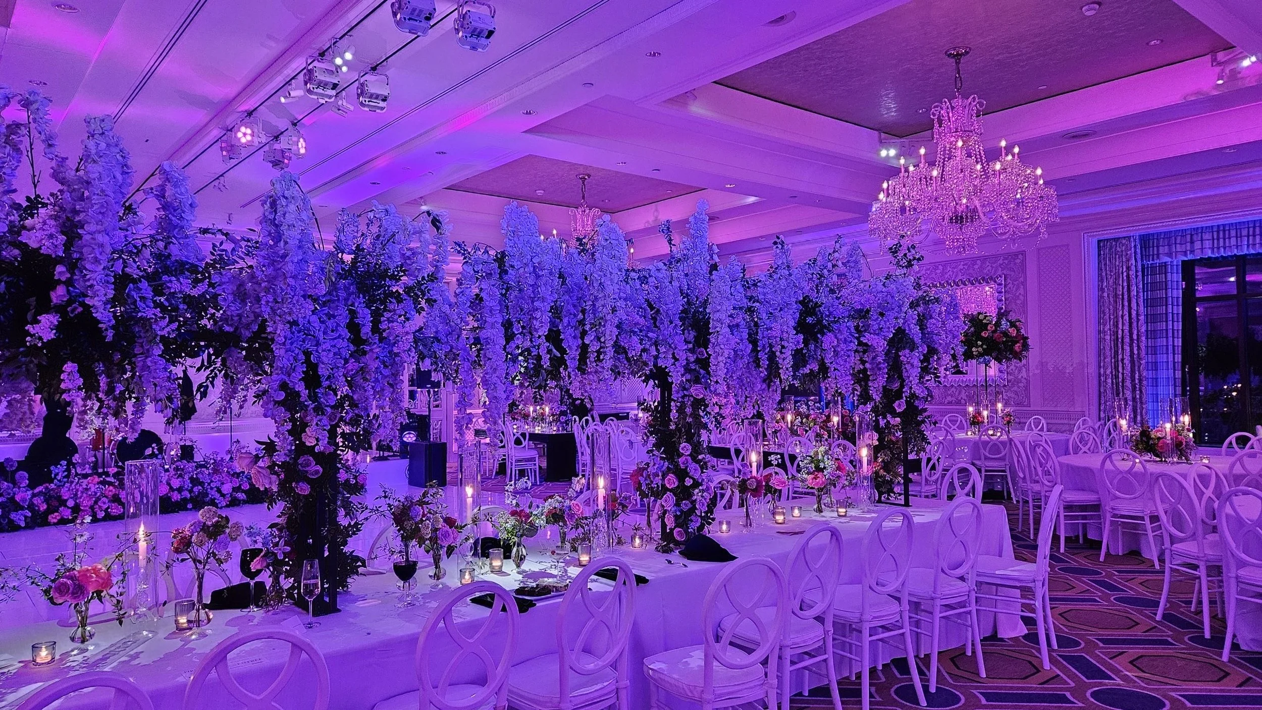Elegant banquet hall decorated with purple and pink flowers, tall floral centerpieces, candelabras, and a large chandelier, set for a formal event or wedding reception.