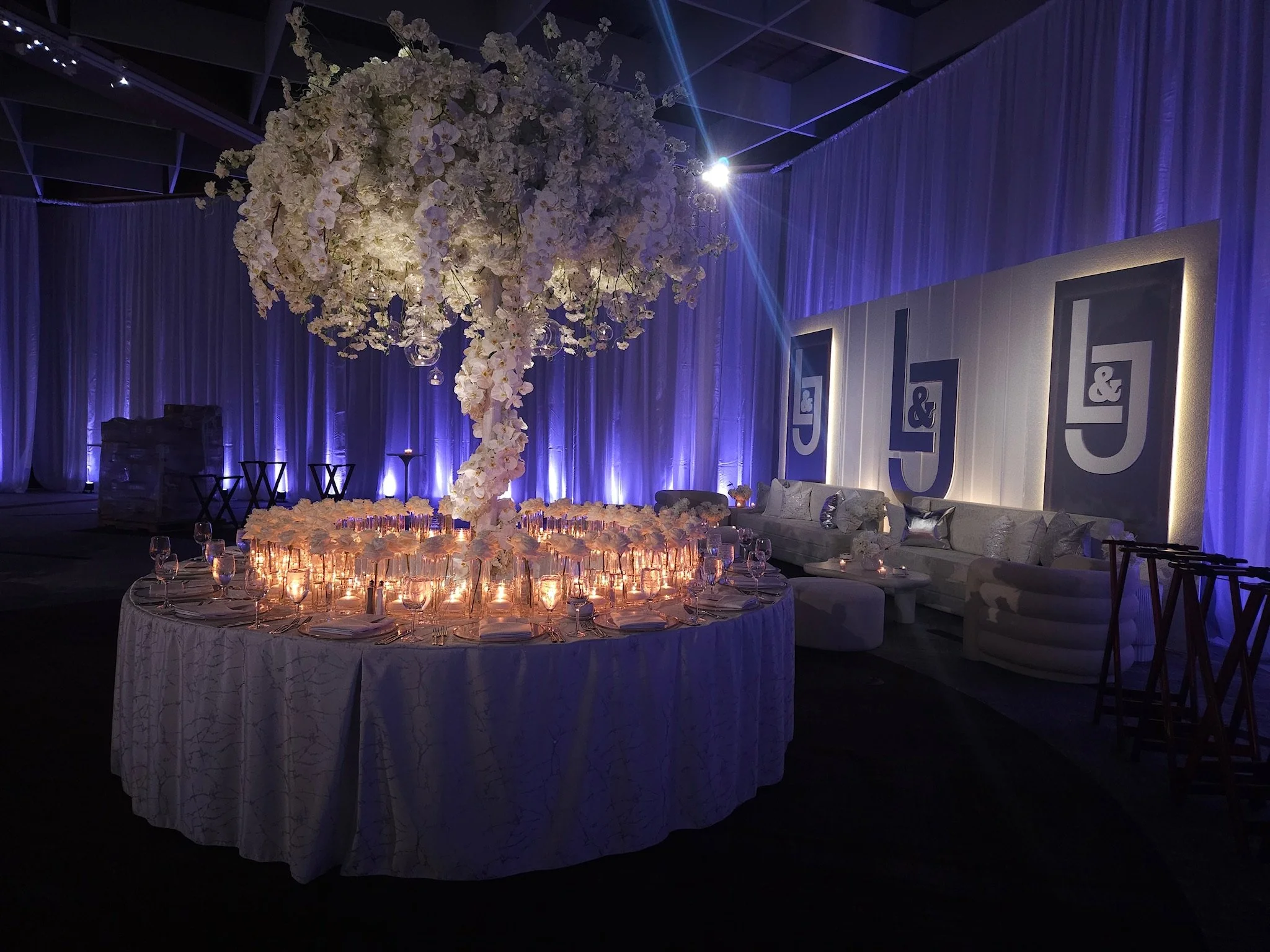 Elegant event setup with a large floral centerpiece chandelier over a round table, illuminated with warm candlelight, surrounded by glassware and table settings, with a white and purple color scheme and a seating area with couches and artwork with th