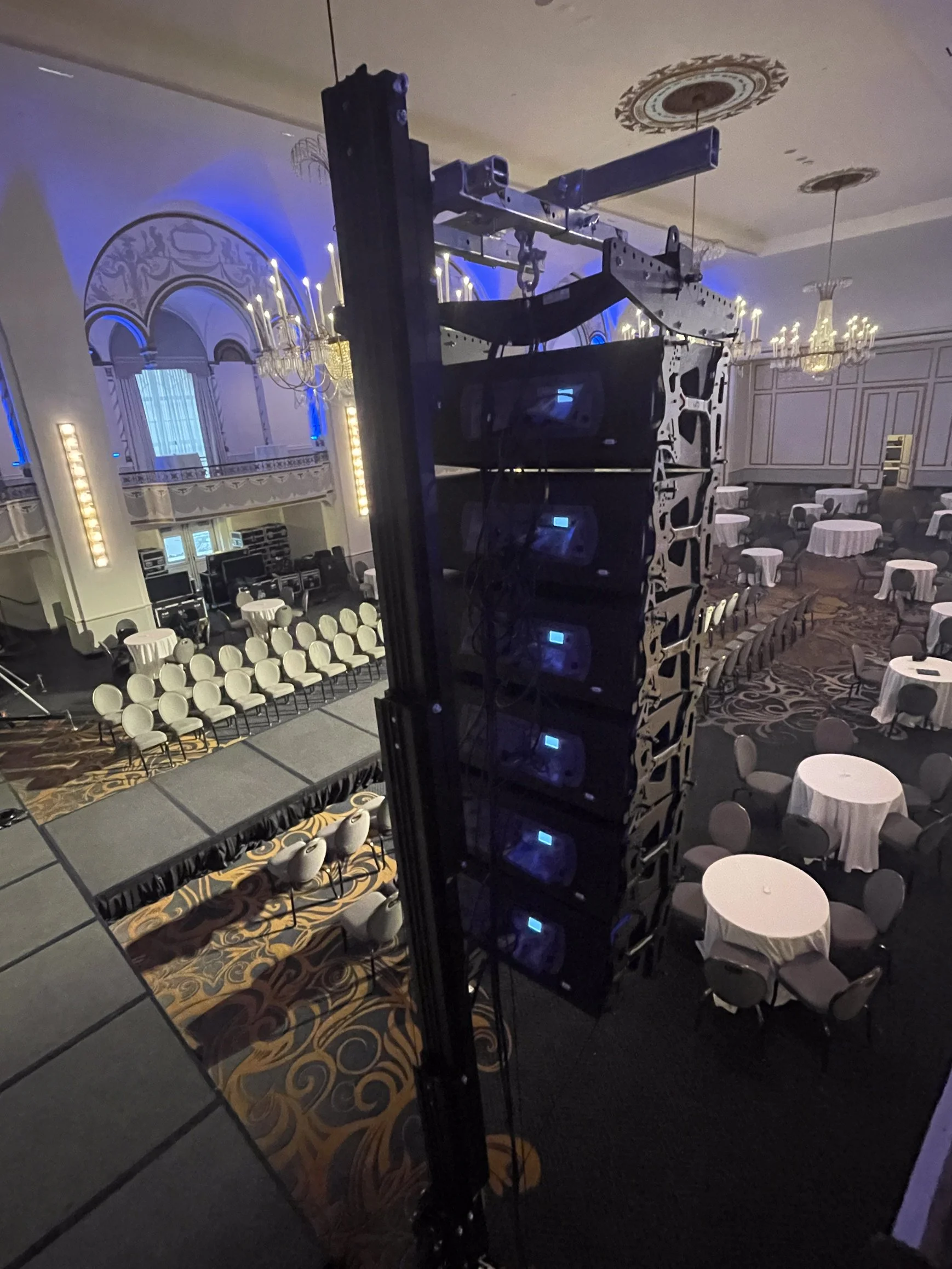 View of a stage setup with large speakers hung from a rig in an elegant banquet hall with chandeliers and round tables.