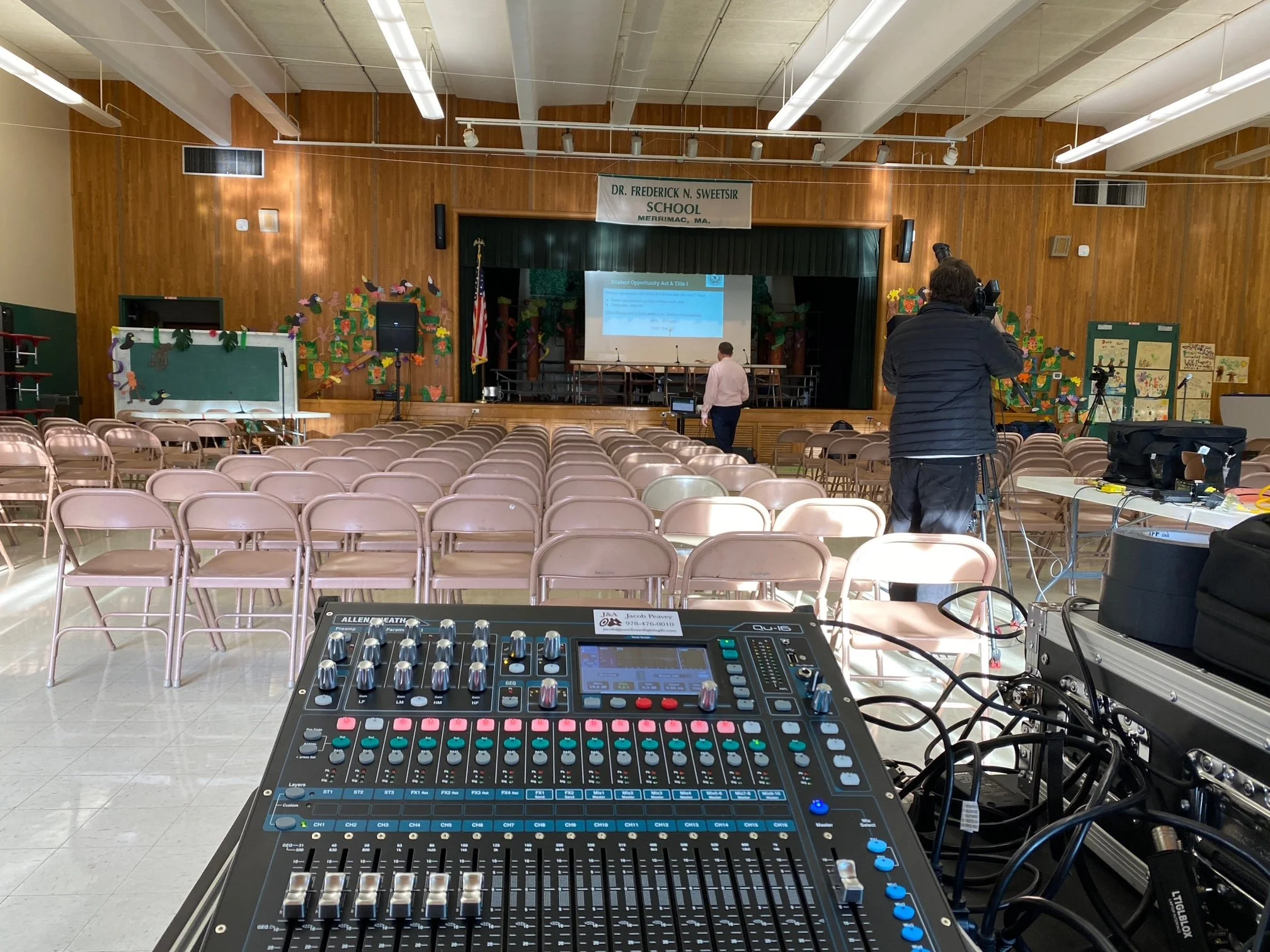 An auditorium with rows of empty chairs and a stage at the front, decorated with colorful paper crafts and artwork on the walls. A person is setting up equipment near the stage, and a camera operator is filming.