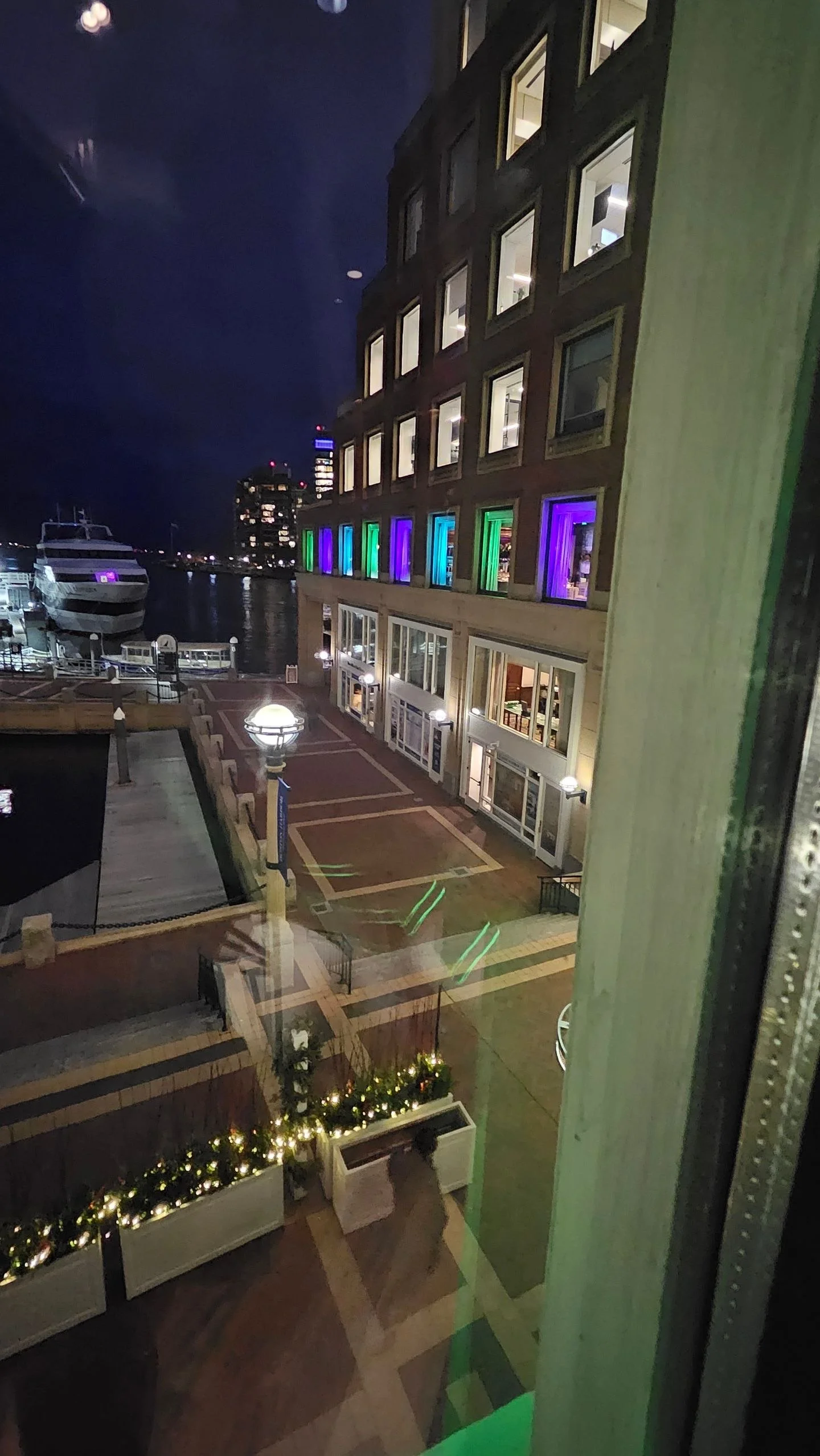 Nighttime view of a multi-story building with large windows, some illuminated inside, and colorful neon lights on some of the windows. A waterfront harbor with boats and yachts is visible in the background. The scene includes a pathway with lampposts