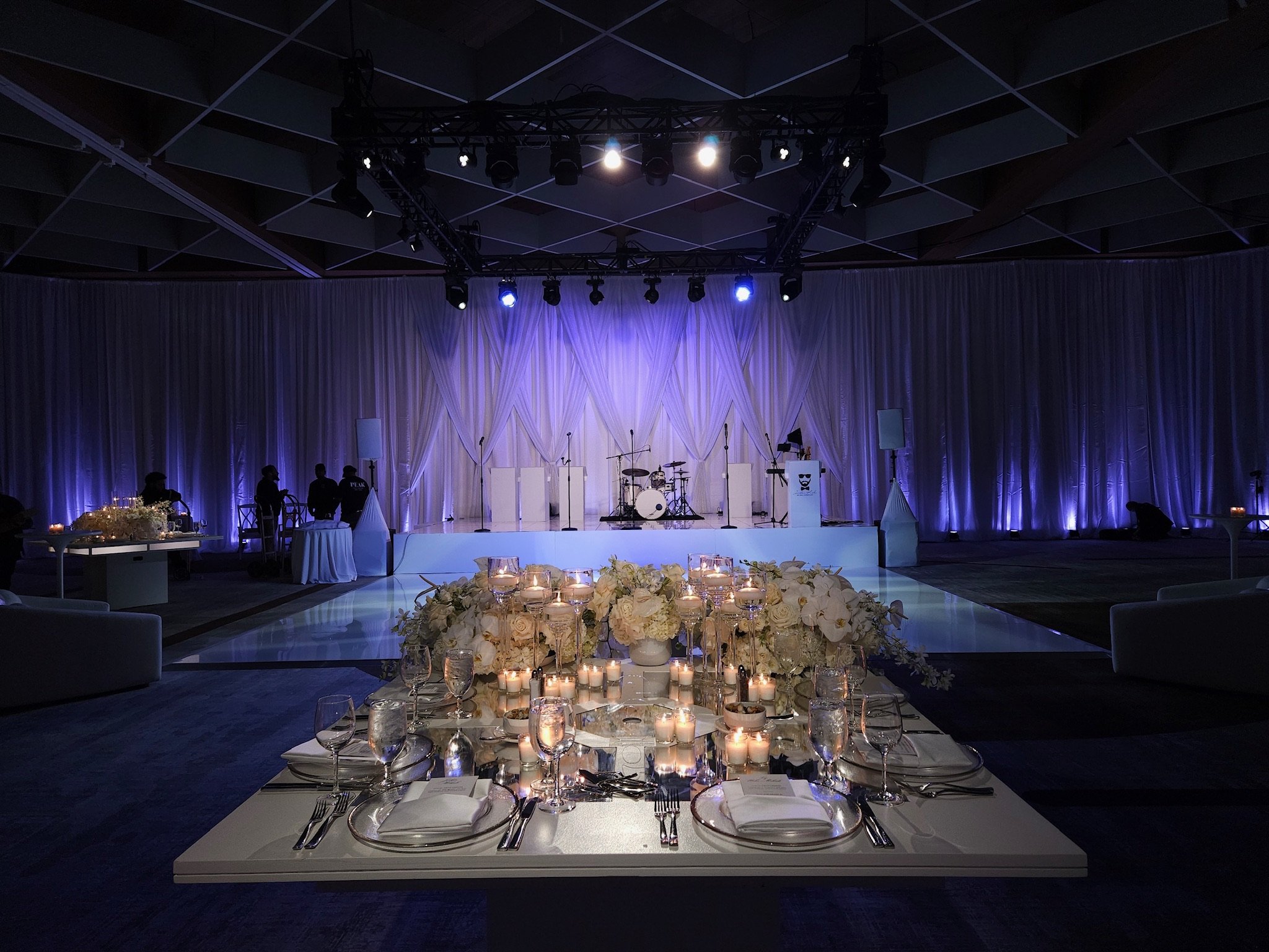 Elegant event space with a stage, musical instruments, and purple lighting, decorated with white flowers and candles on a dining table.
