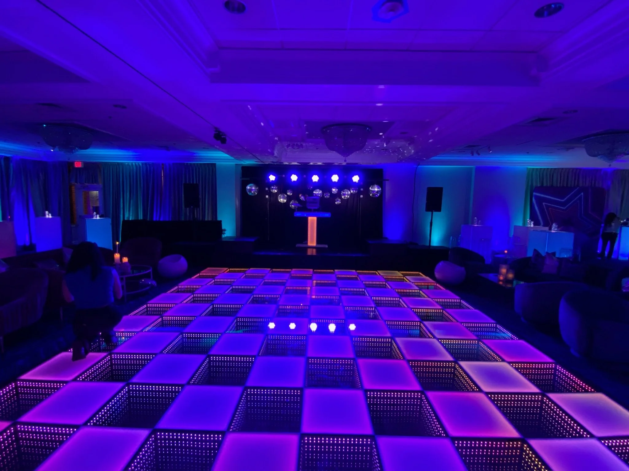 A dimly lit room with a checkered dance floor illuminated in purple and pink lights, a stage with a DJ booth, and scattered seating areas with couches and tables, set for a nightlife or party event.
