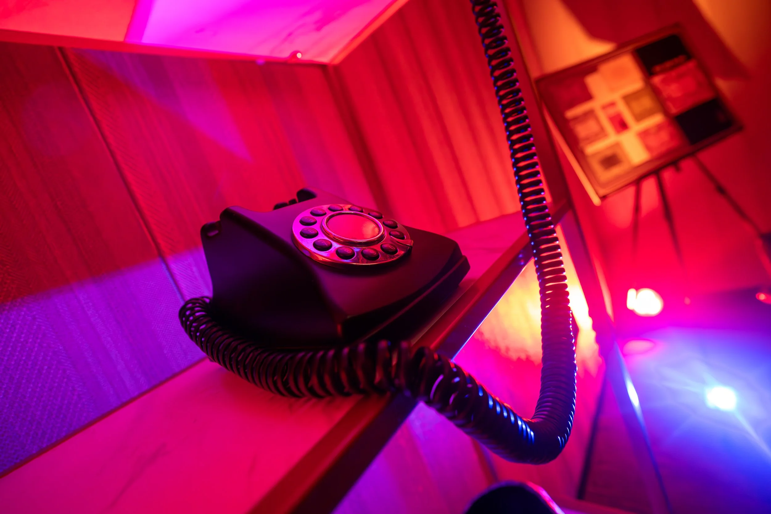 A vintage rotary phone resting on a wooden table, illuminated by colorful pink, purple, and blue neon lights in a dim room with a wooden wall, a framed picture, and a tripod in the background.